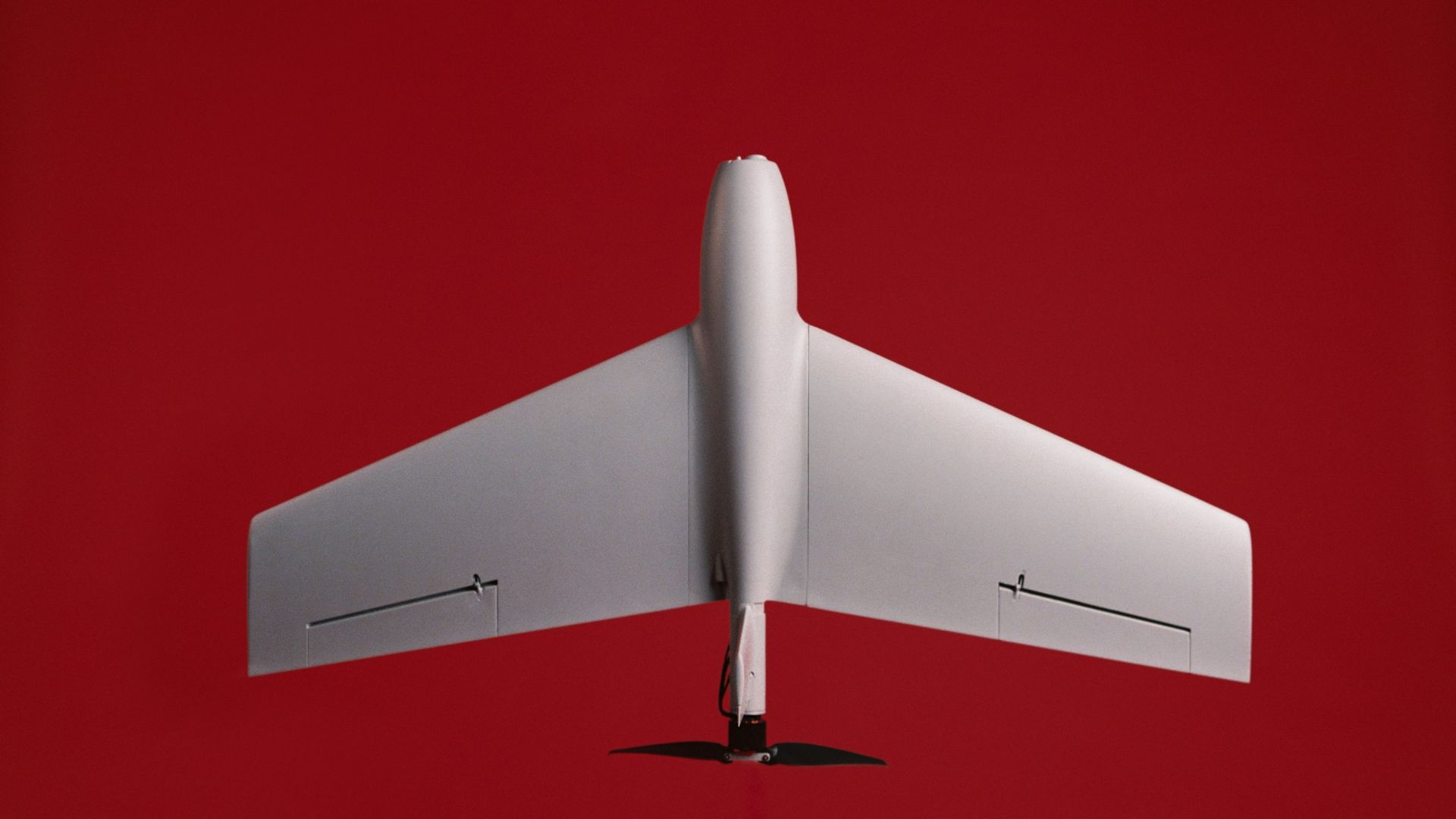 Top-down view of a white model airplane with a tall fuselage and straight wings, set against a solid red background. A small black propeller sits below the fuselage.