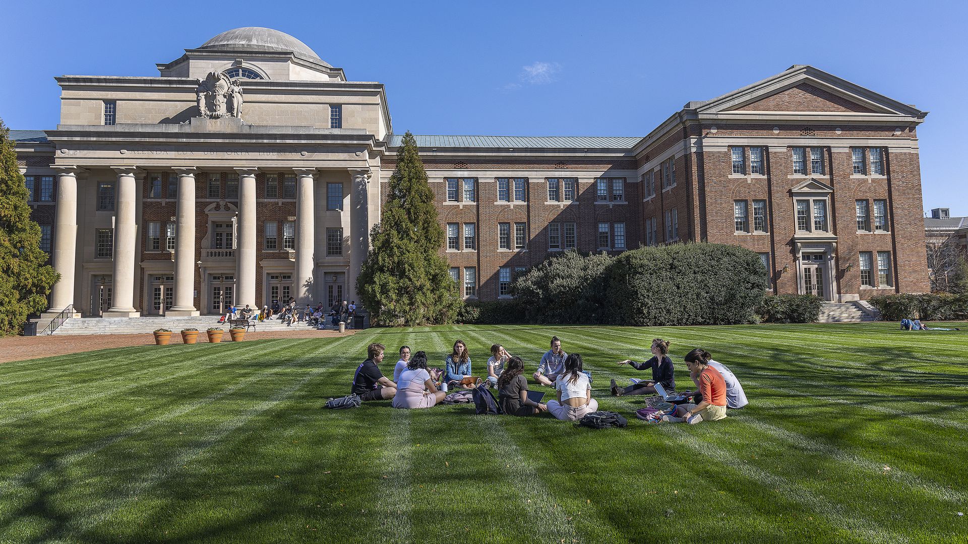 An academic building with a large dome, classical columns, and brick wings, under a clear blue sky; students sit on a well-kept green lawn near the entrance.