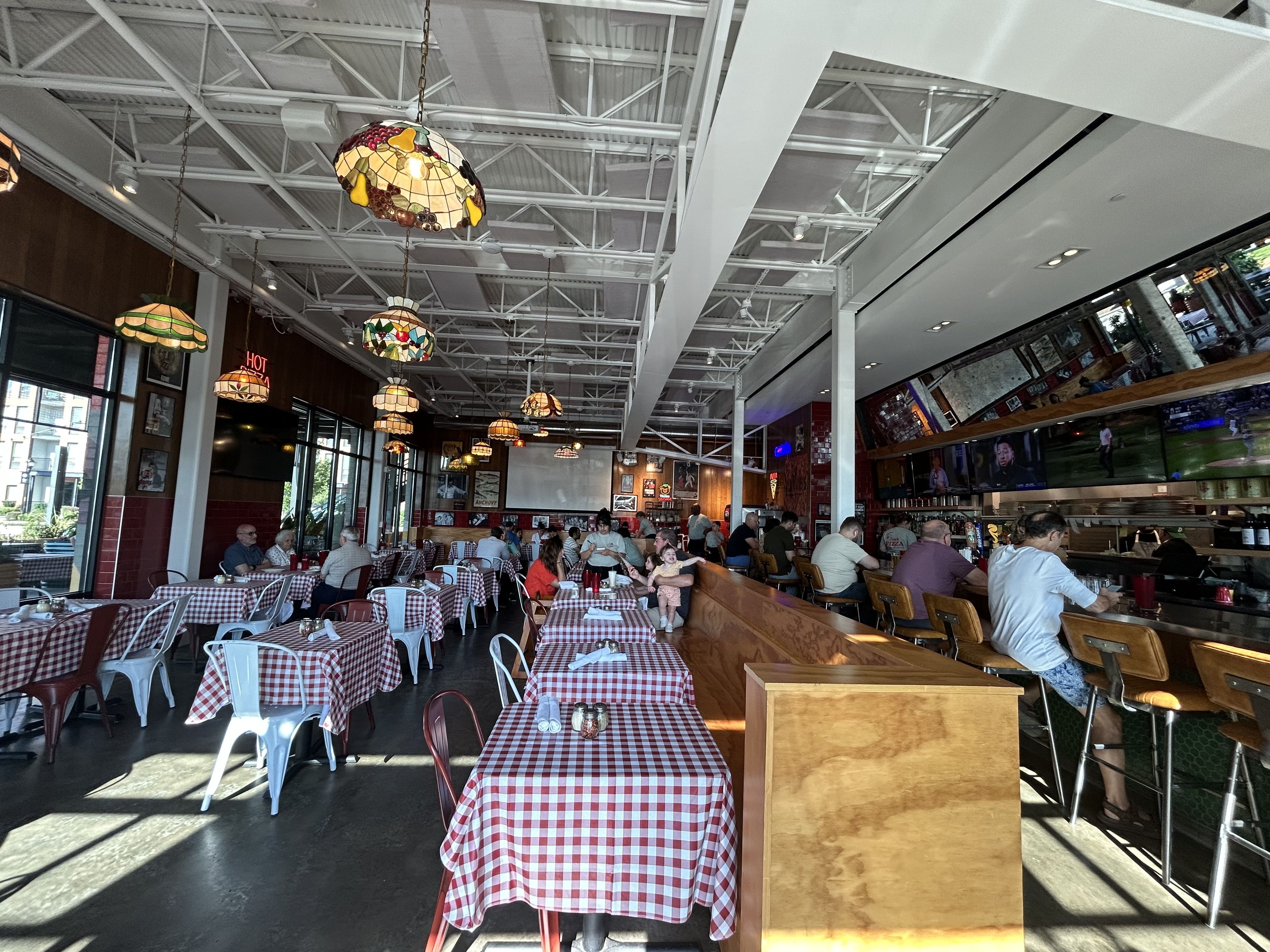 Interior of a busy restaurant with red and white checkered tablecloths, stained glass pendant lamps, wooden booths, metal chairs, and multiple TVs above the bar showing sports.