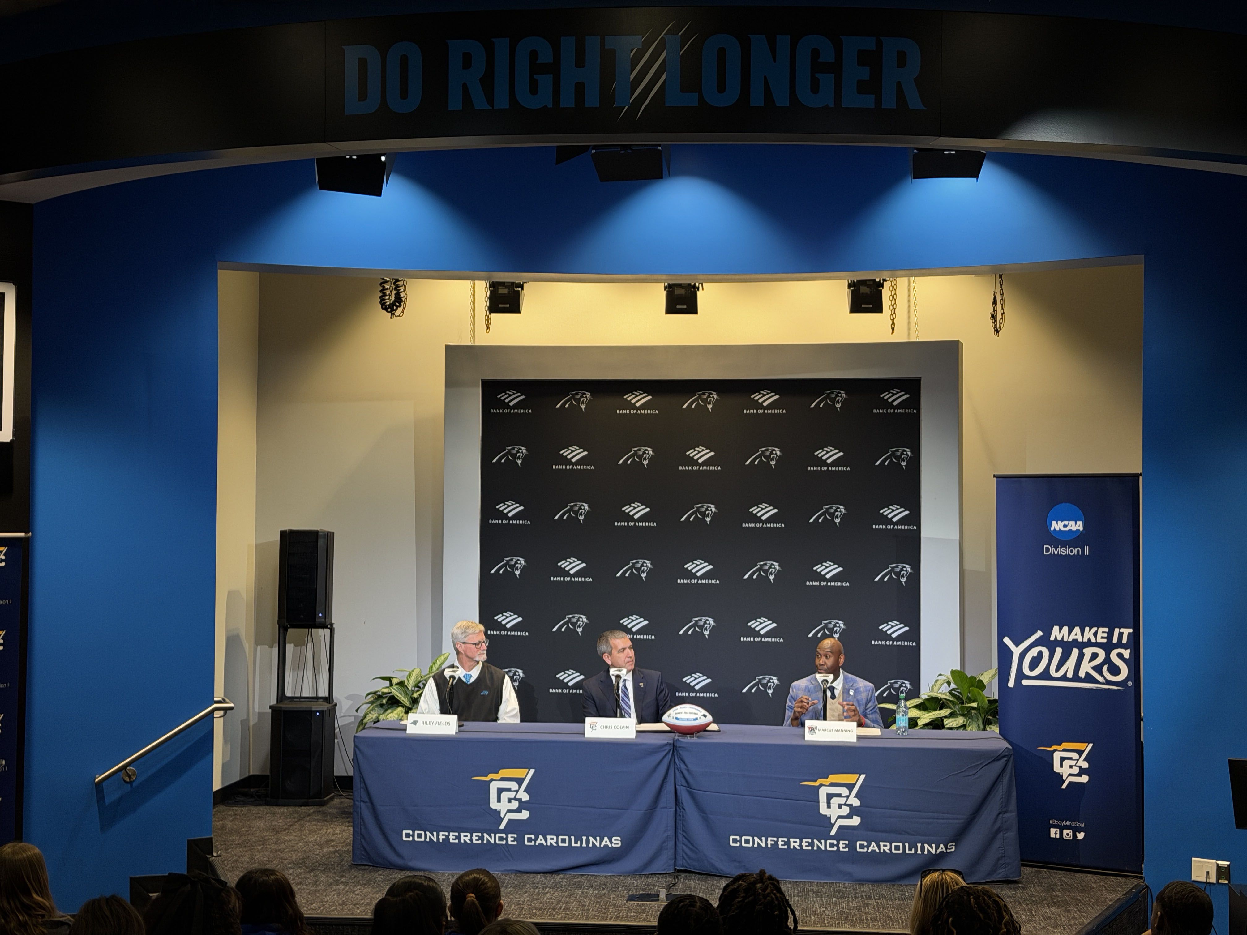 Three men seated at a table with Conference Carolinas banners, a football, and signage behind. Above them a sign reads "DO RIGHT LONGER" in blue letters.