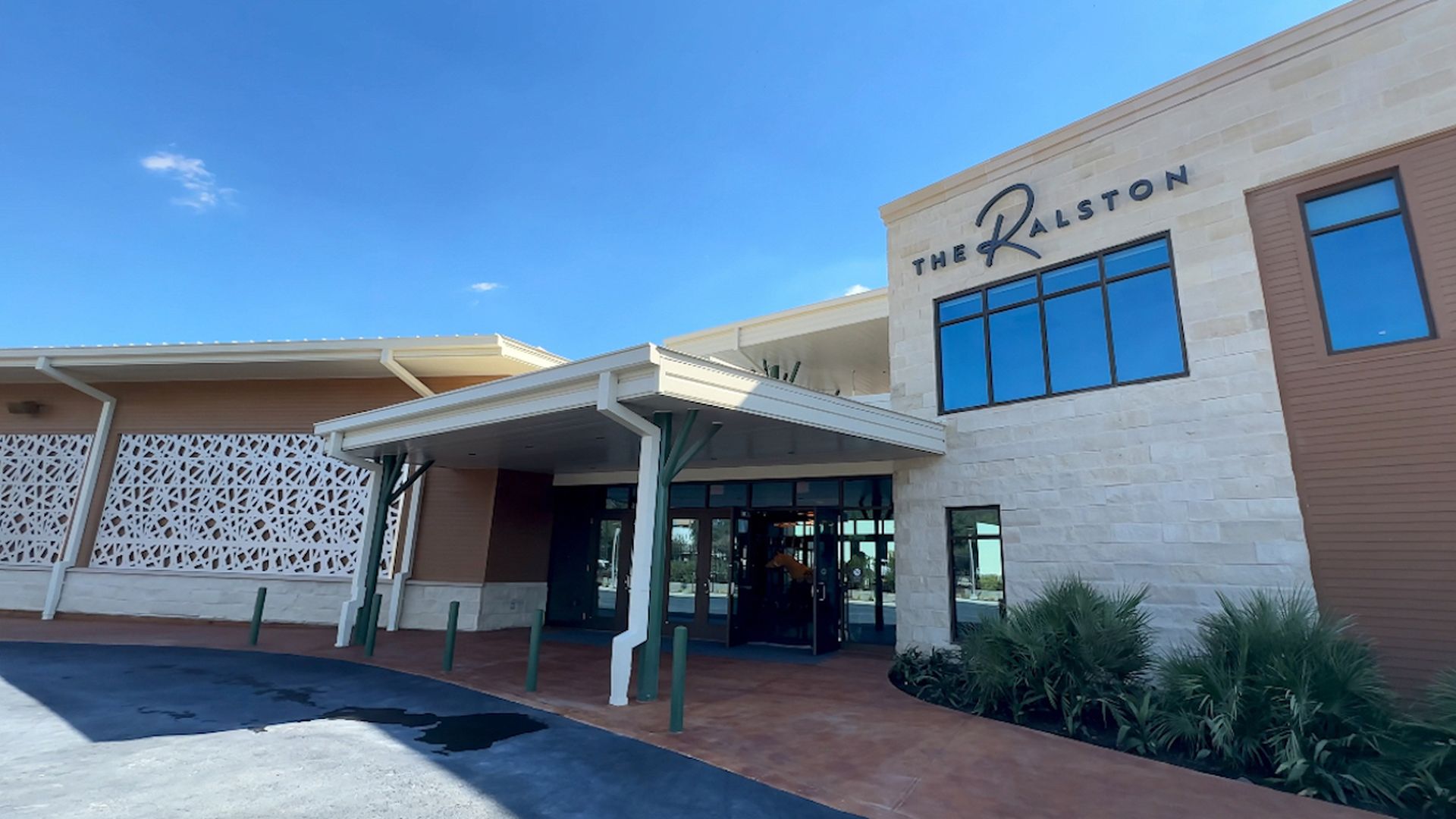 Entrance of The Ralston building with beige stone and brown siding under a bright blue sky, featuring a covered walkway, green landscaping, and clear glass windows.