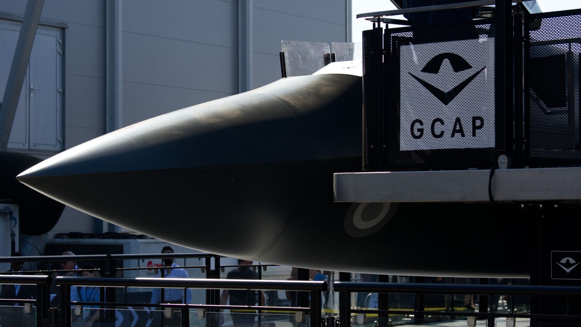 The nose of a life-size model aircraft peeks around from an observer stand. It's sunny out.