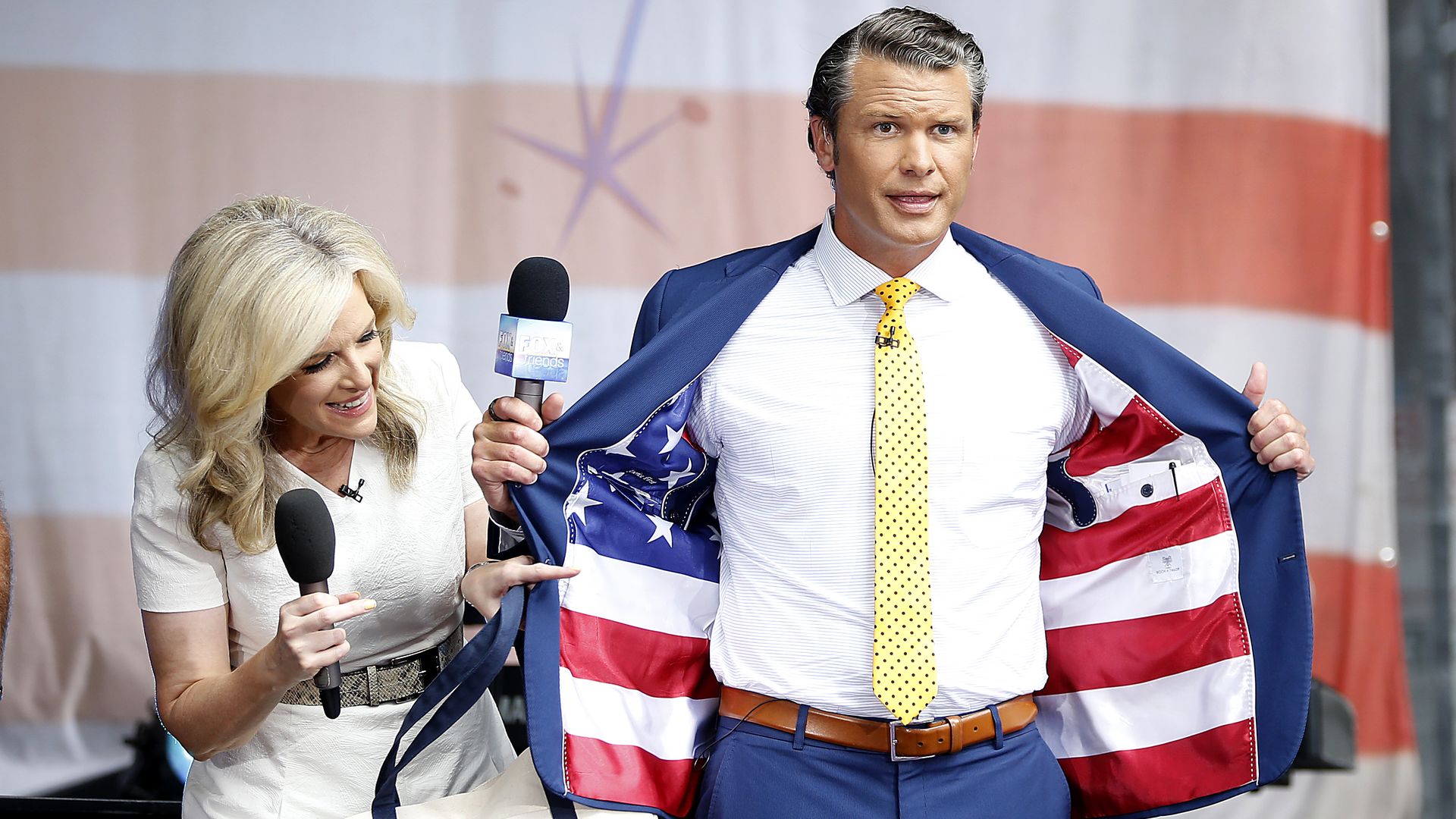 A woman and man stand on stage holding mics. A large American flag is the backdrop. The man is in a blue suit with yellow tie.