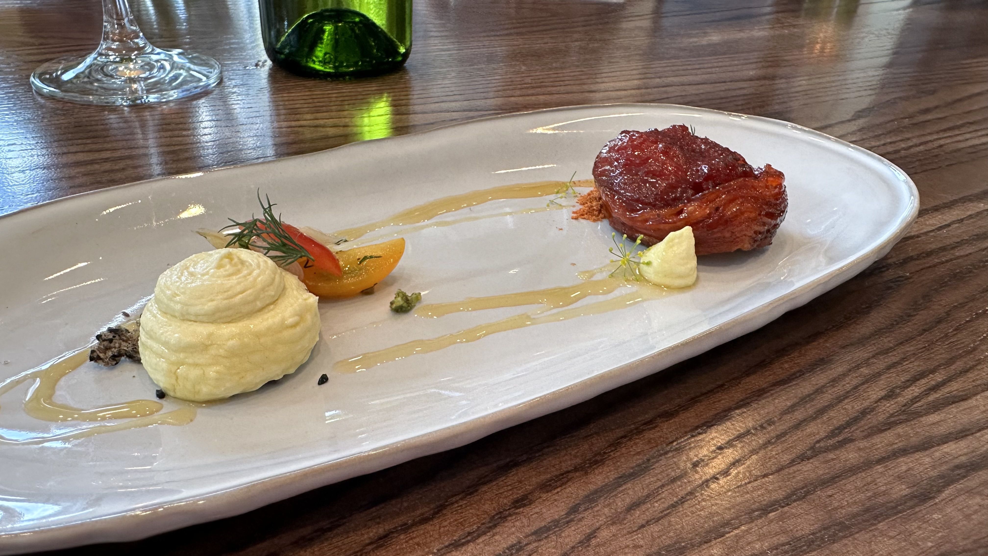 A long white plate is filled with a light yellow dollop of cream and a red tomato pastry.
