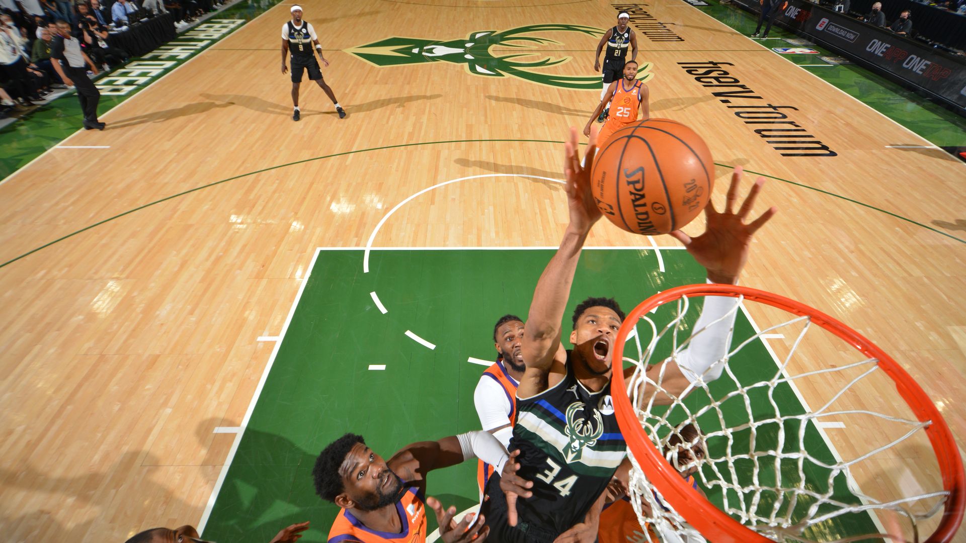 Giannis Antetokounmpo, in a green jersey, #34 of the Milwaukee Bucks, jumps to dunk a basketball into a hoop above Phoenix Suns players in orange uniforms.