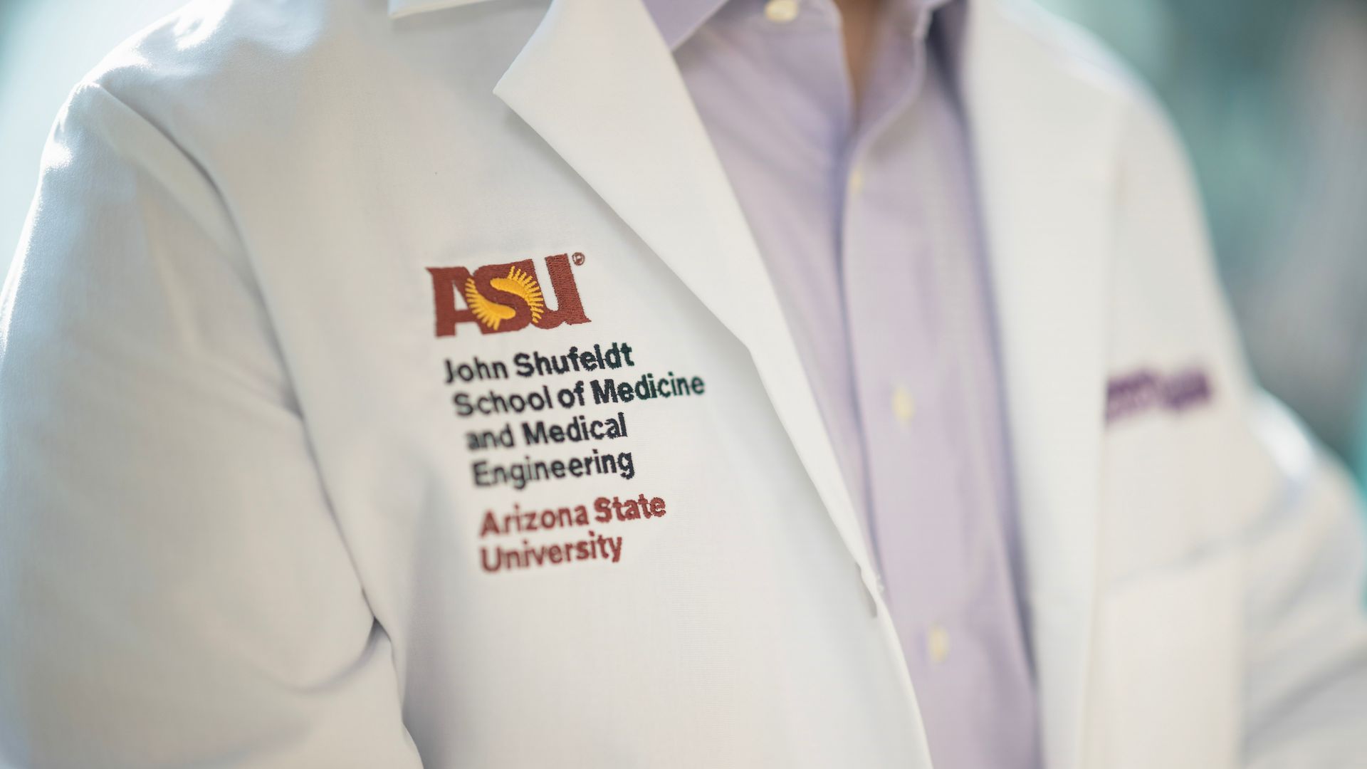 Close-up of a white lab coat with the ASU logo and text "John Shufeldt School of Medicine and Medical Engineering, Arizona State University" embroidered in maroon and gold.