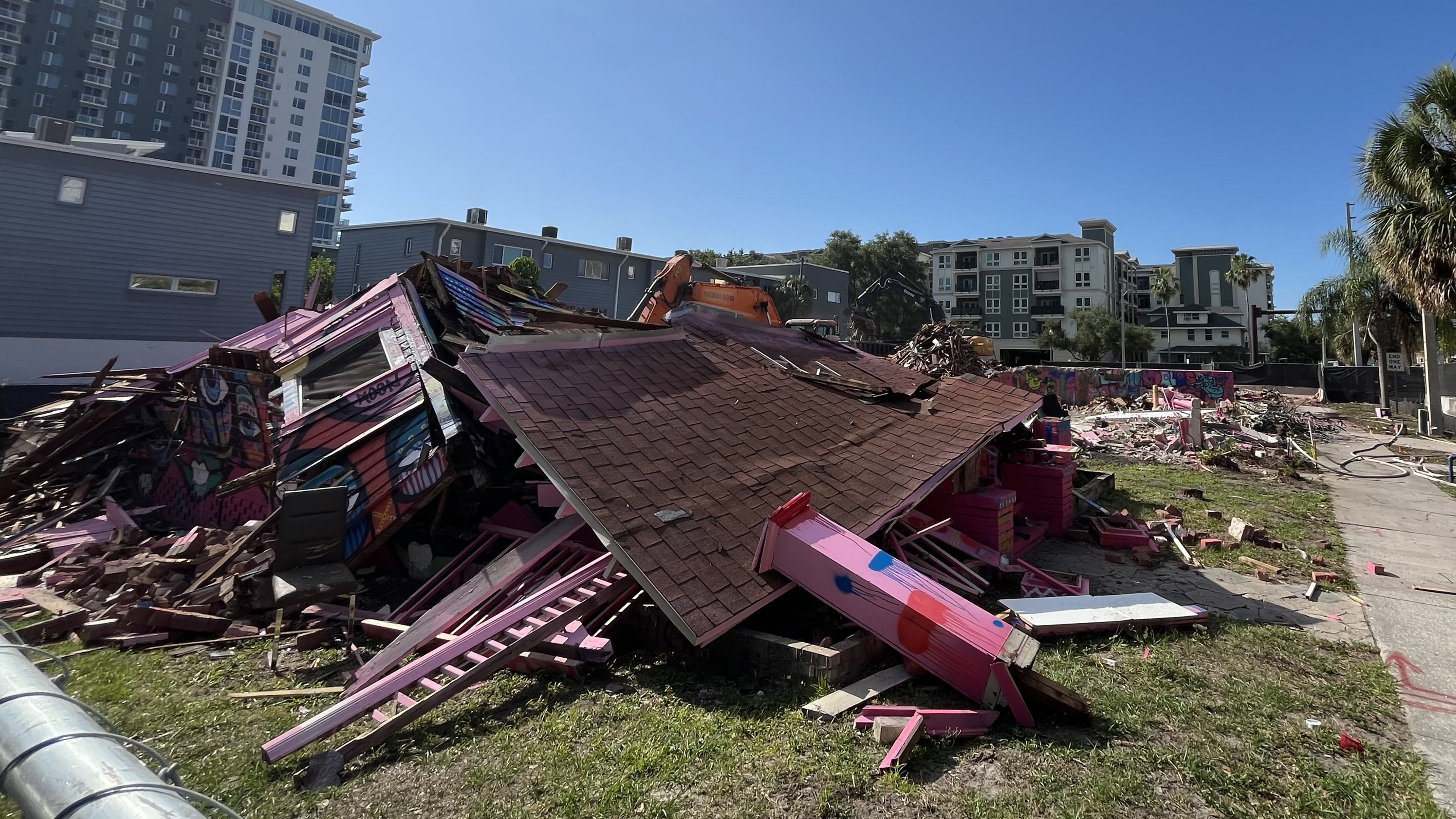 Blue sky over a city demolition site with crushed pink building rubble and an orange excavator, surrounded by gray modern apartments and palm trees in the background.
