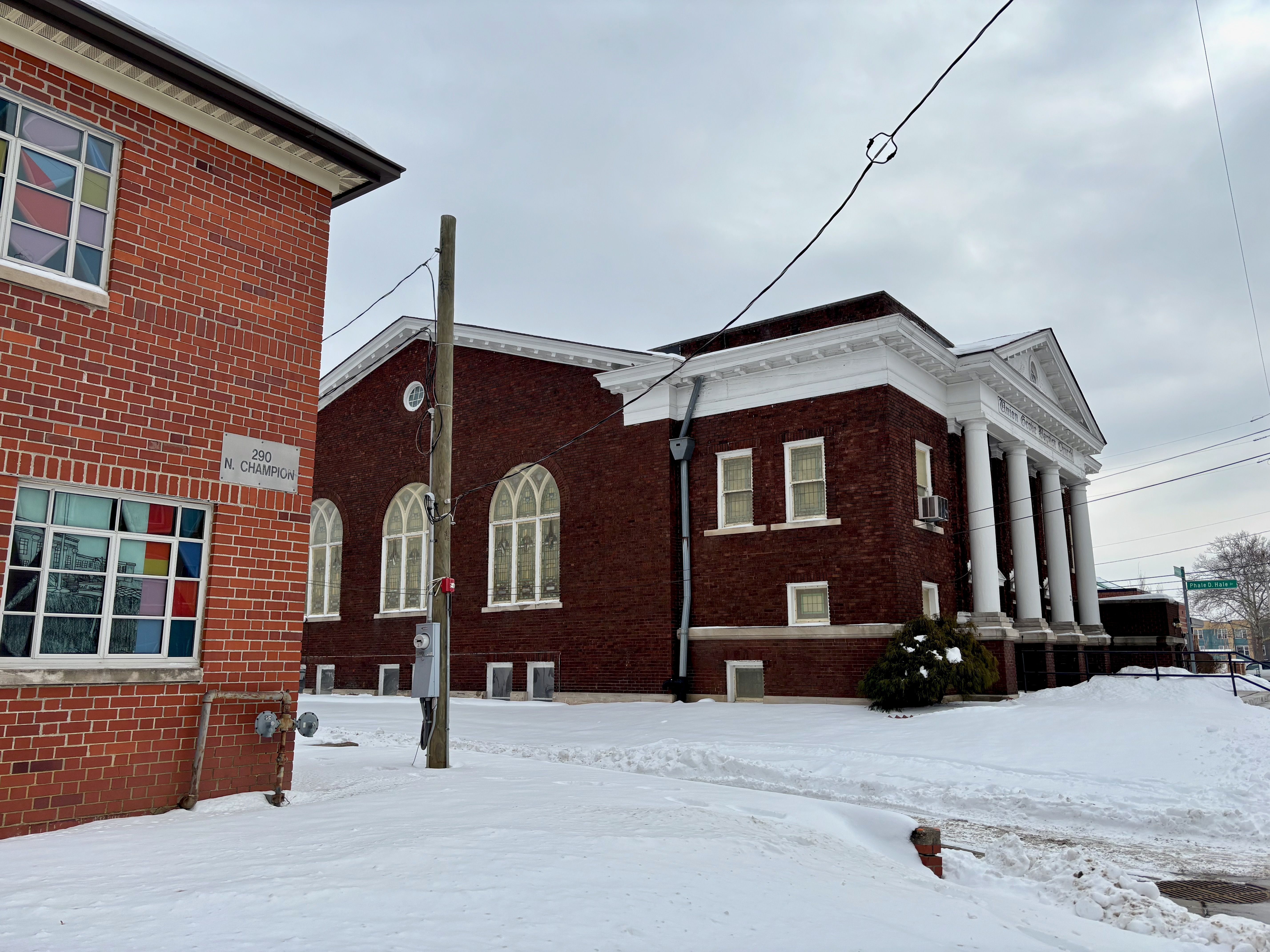 Snow-covered ground with two red brick buildings, one with stained glass windows and the other with white columns, under a cloudy sky in a quiet street corner.