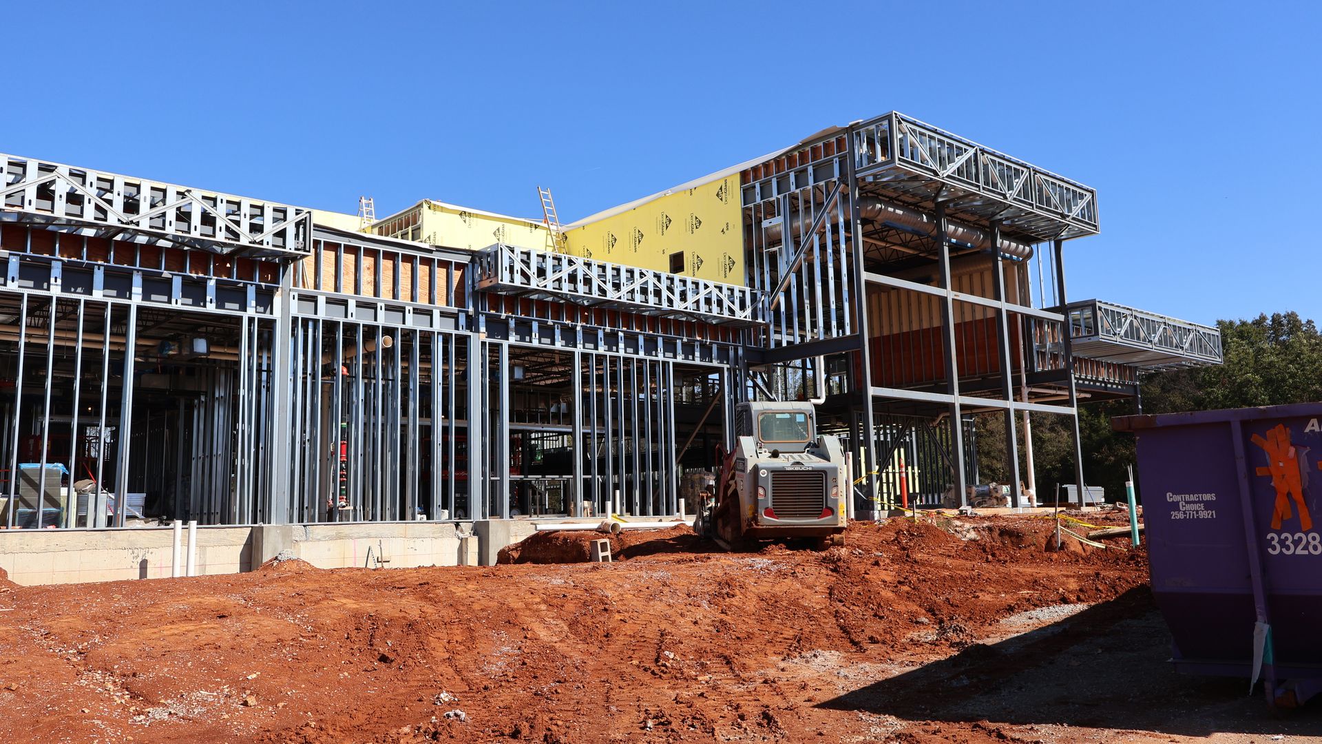 Steel frame structure of a building under construction on red dirt ground, with a small construction vehicle and a purple dumpster on site under a clear blue sky.