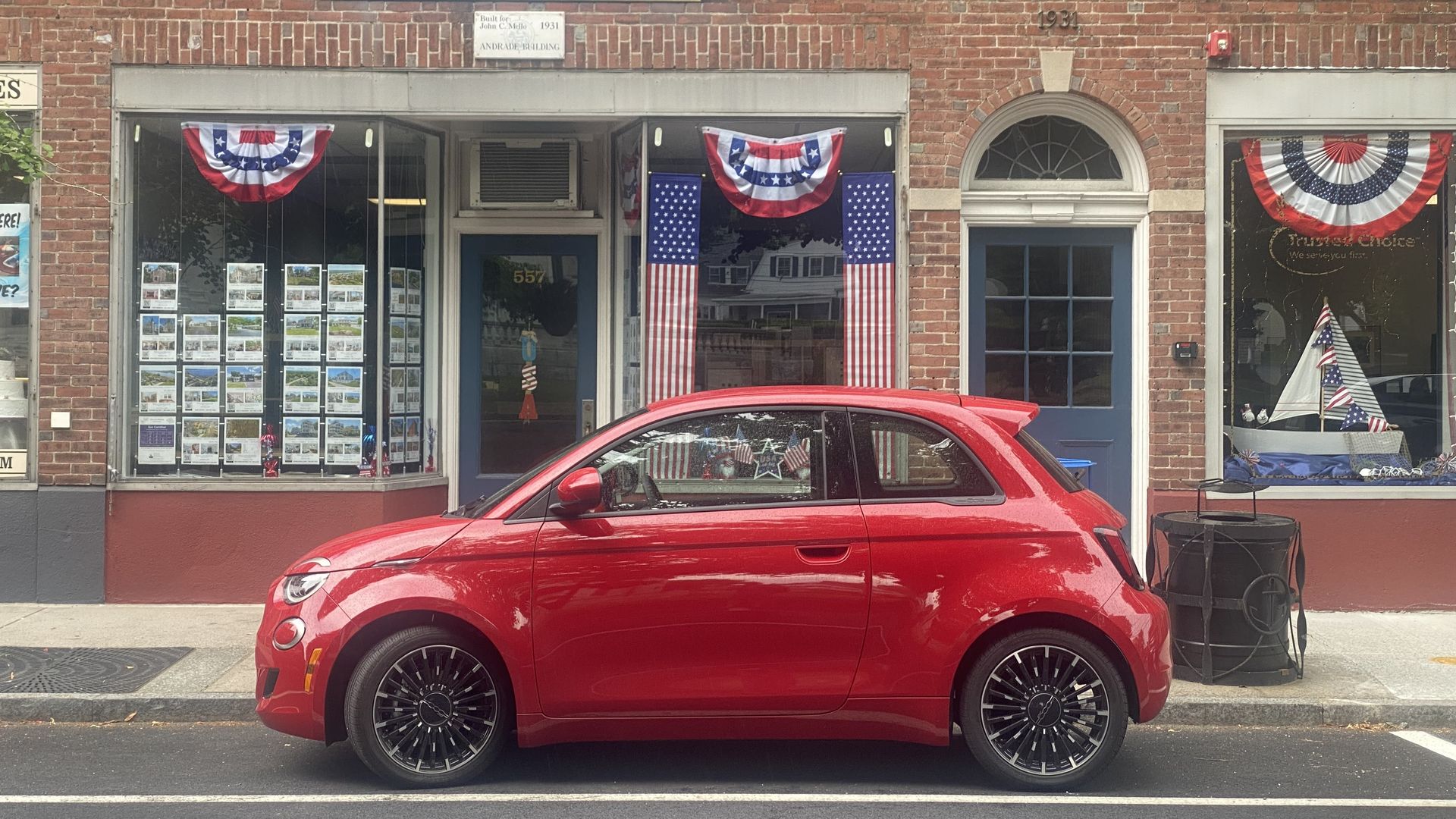 A photo of a bright red Fiat 500e electric hatchback, parked on the street in front of a real estate storefront decorated with American flags and bunting.
