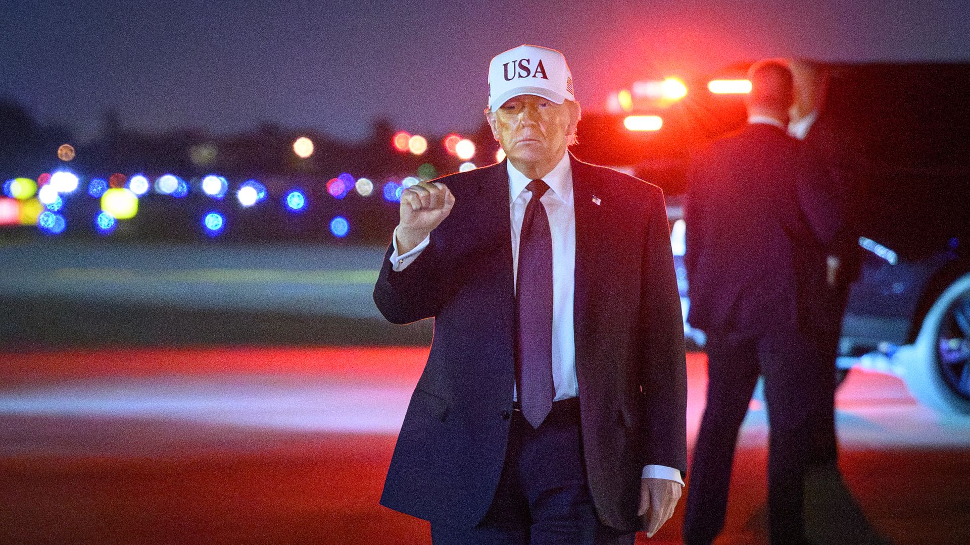 President Trump — wearing a dark suit, a white collared shirt, a dark tie, and an American flag pin and a white "USA" cap — holds his fist up as he walks.