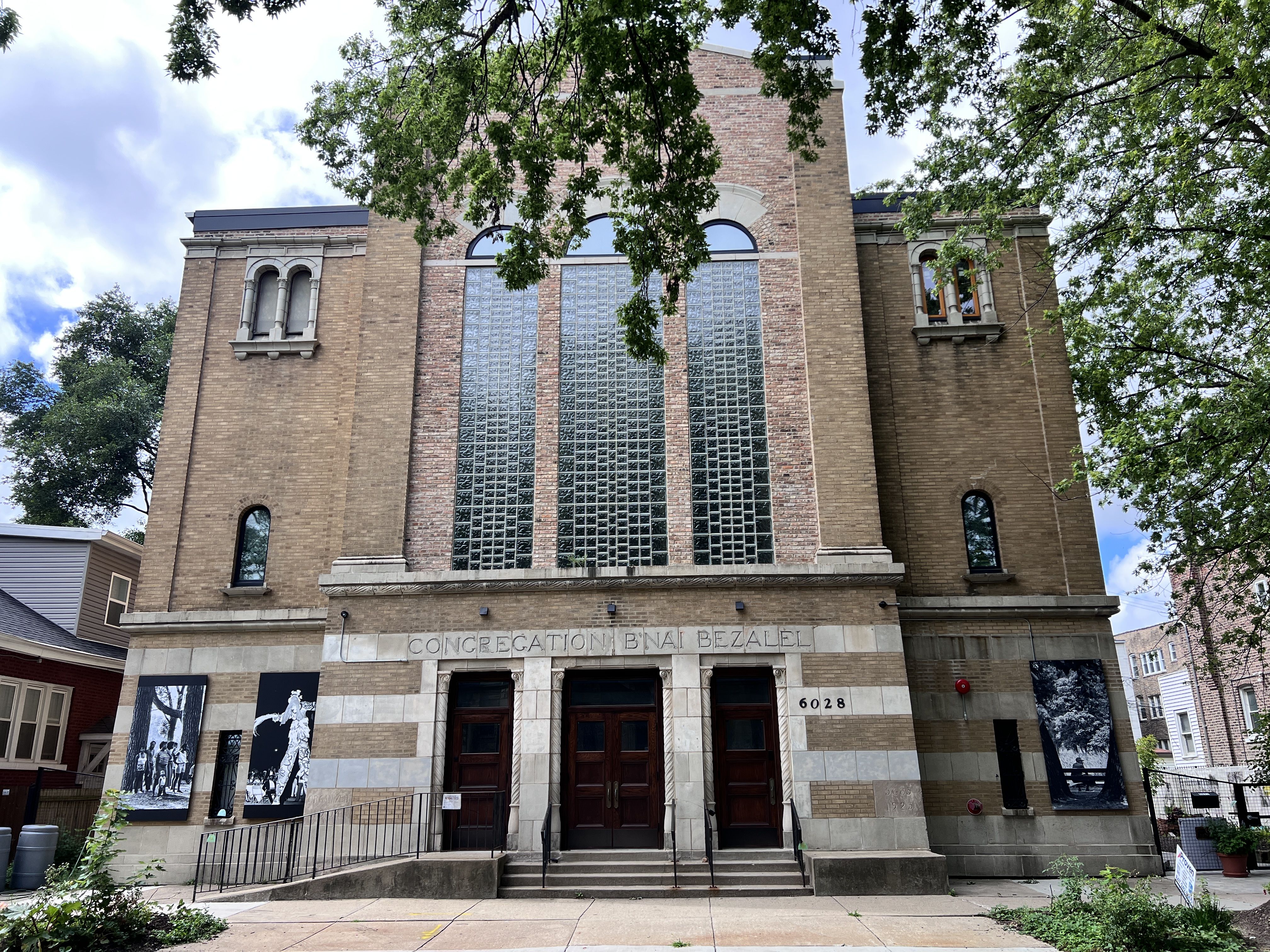 Brick synagogue with tall glass-block center, arched windows, and decorative stonework; front entrance with double doors and steps. Framed by green trees; sign reads "Congregation B'nai Bezalel".