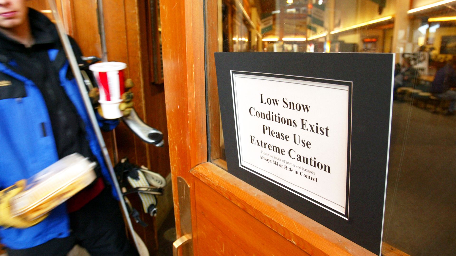 Sign on a wooden door reading, "Low Snow Conditions Exist Please Use Extreme Caution." Person in blue jacket holding skis, food, and drink entering a lodge interior.