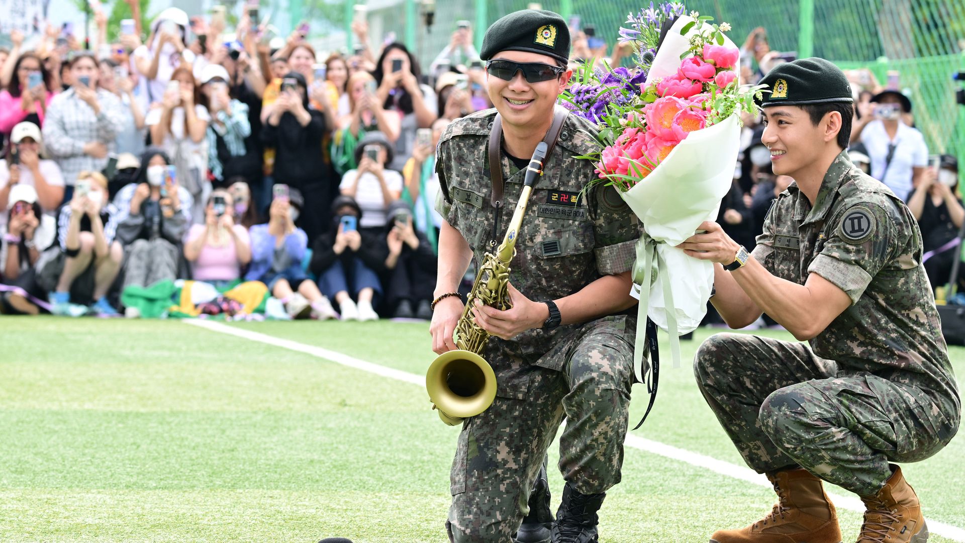 Two soldiers in camouflage uniforms kneel on a field, one smiling with sunglasses holding a saxophone, the other presenting a colorful bouquet of flowers, with a crowd taking photos in the background.