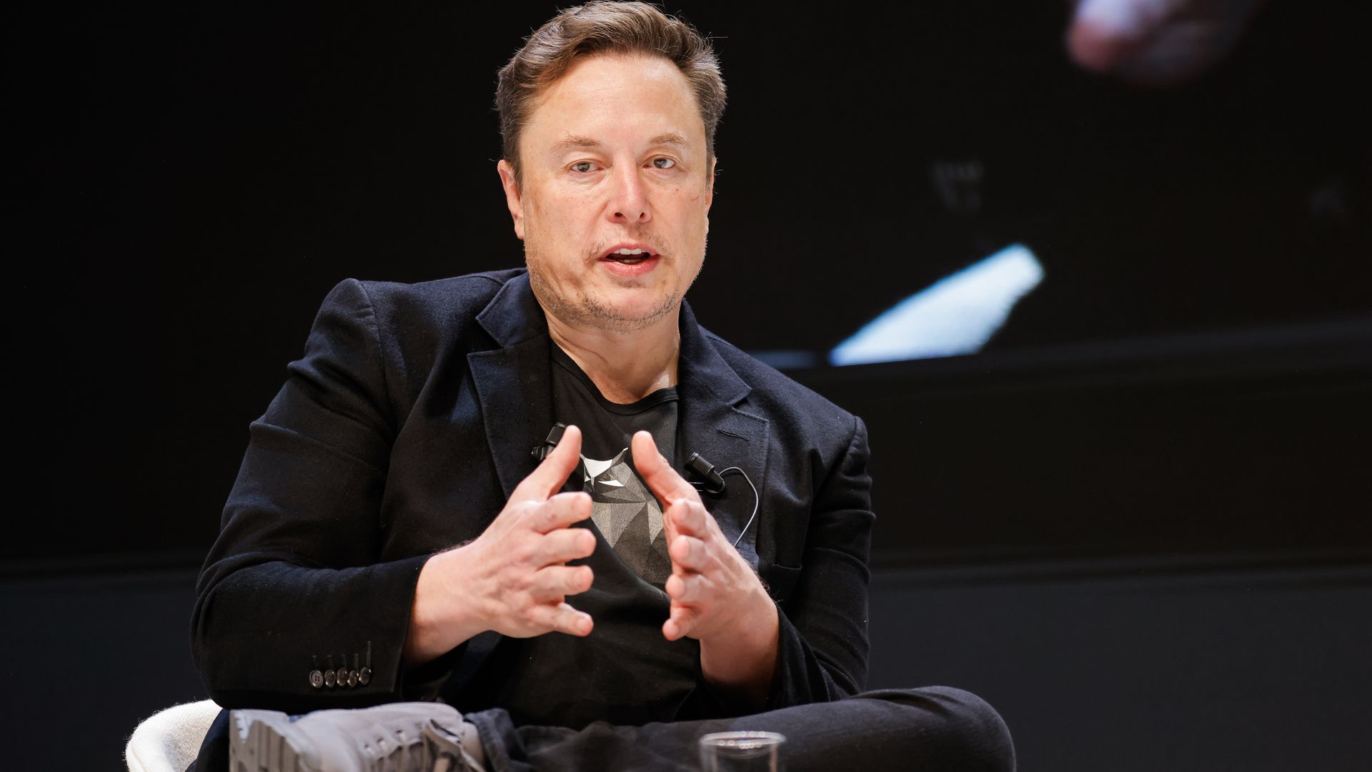 Elon Musk looks into the camera and gestures with his hands close to each other in front of a black background
