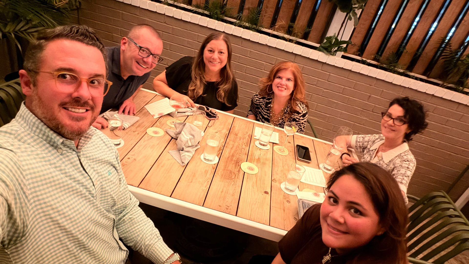 Five friends at an outdoor restaurant table pose for a selfie. Foreground man with glasses smiles; others include one man and two women in the background plus a woman at the right, all with drinks.