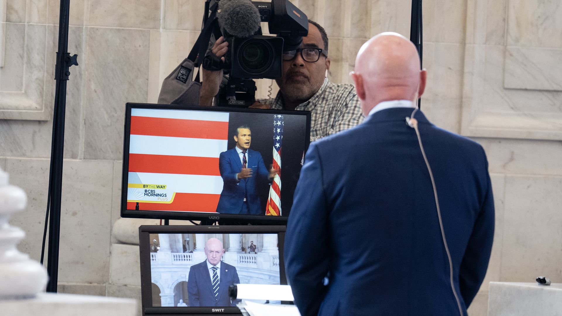 A bald man in a blue suit with a microphone on his back stands in front of two screens, one showing an American flag, the other showing a man in a suit in a marble building.