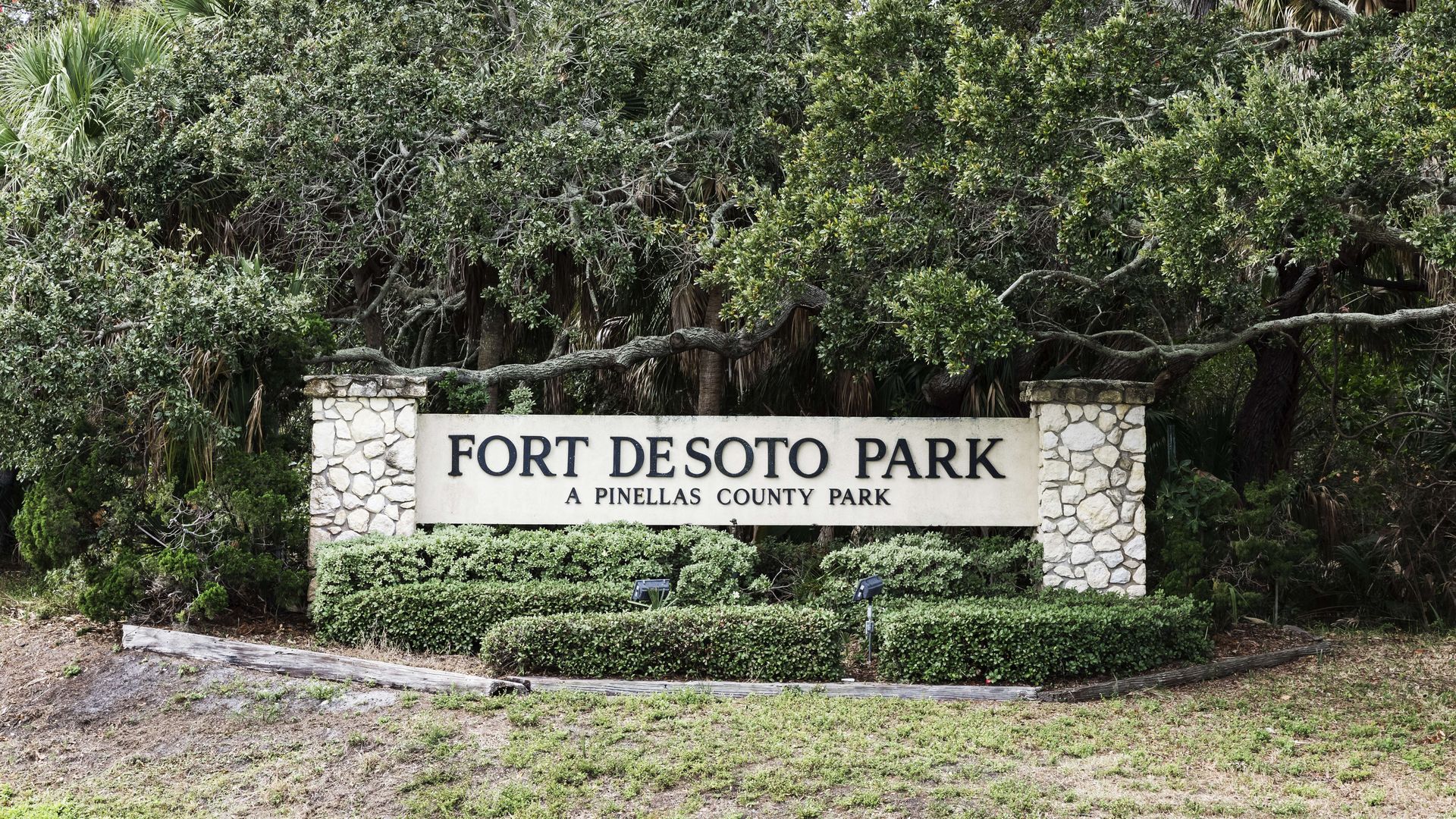 Fort De Soto Park entrance sign: a beige panel with bold black lettering reading 'FORT DESOTO PARK' and 'A PINELLAS COUNTY PARK', framed by stone pillars and surrounded by lush green shrubs and trees.