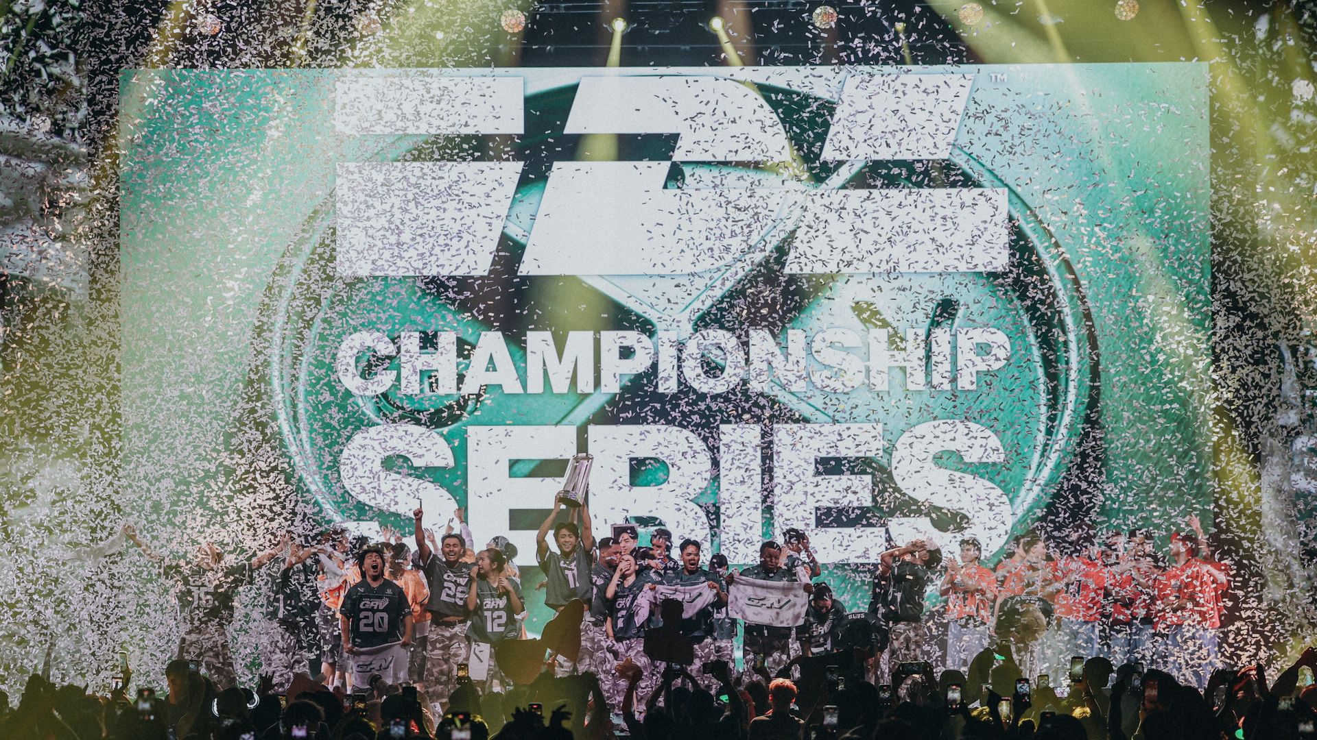 Large crowd cheers on a stage as confetti rains down; a green banner behind players reads "CHAMPIONSHIP" with a logo, while fans photograph the victory from the foreground.