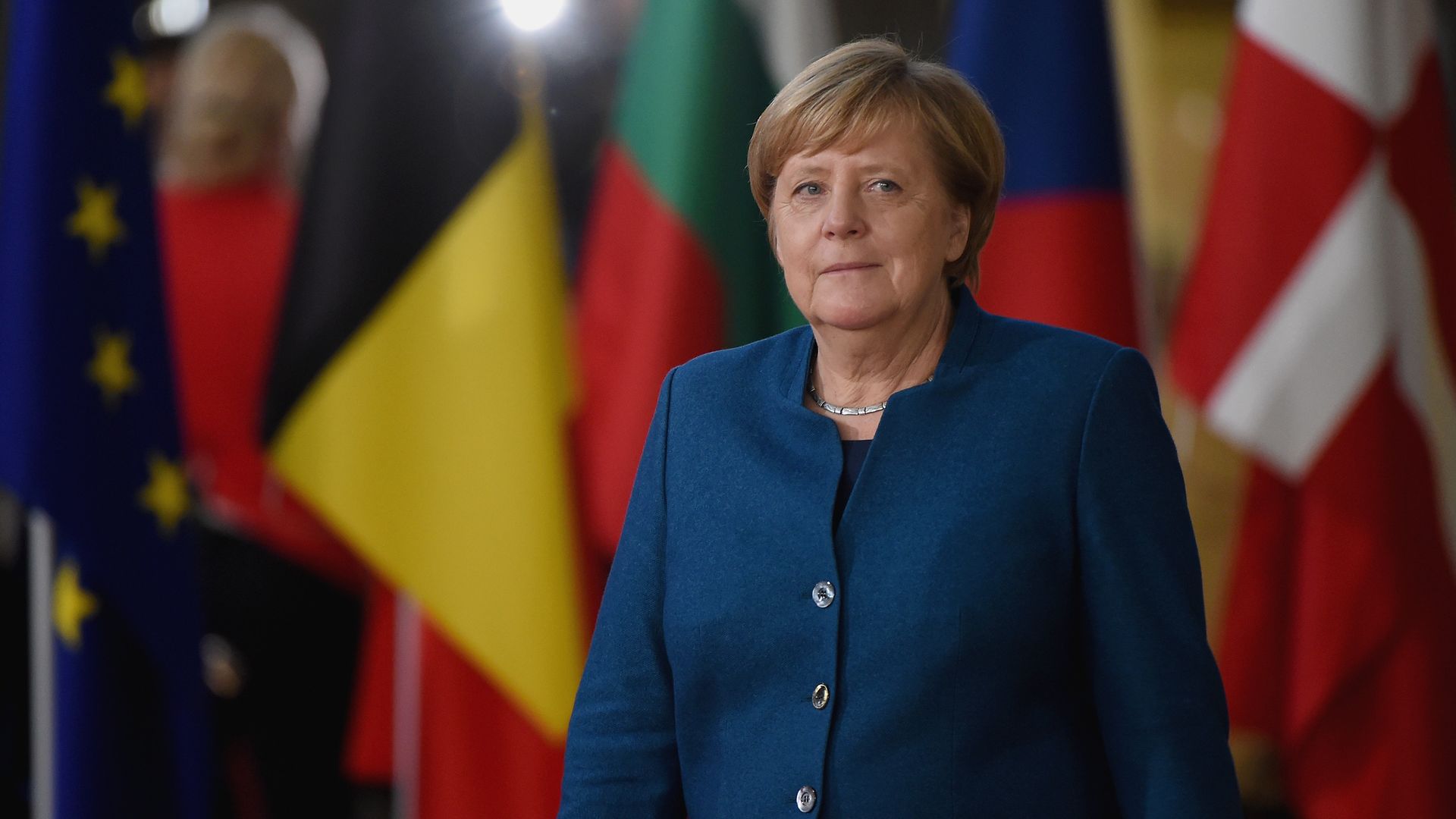 Angela Merkel standing in front of european flags in a blue pantsuit. 