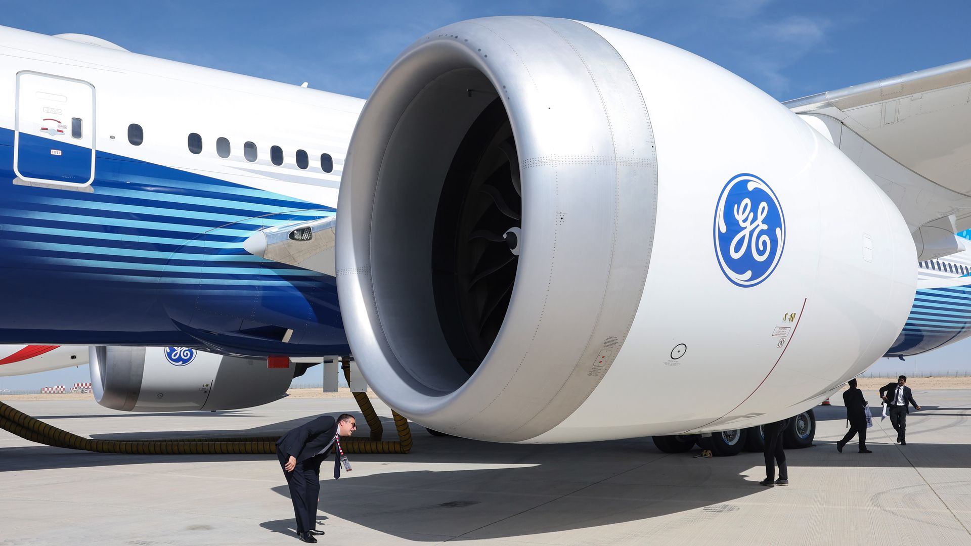Close-up of a large white airplane engine with GE logo, with a man in a suit leaning forward inspecting it on a sunny day, and other people in suits walking nearby on runway.