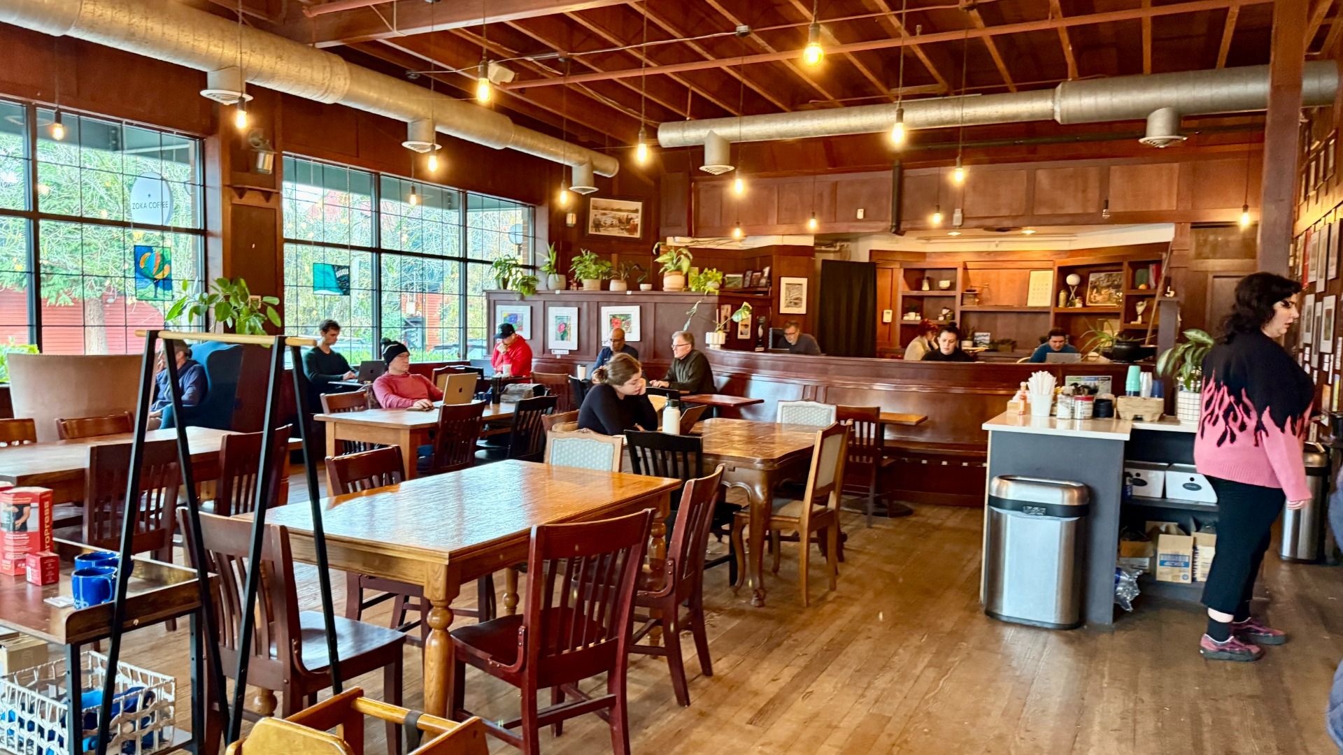 Cozy coffee shop interior with wooden tables, chairs, warm lighting, large windows, several people working on laptops, and a woman in a pink and black sweater standing near the counter.