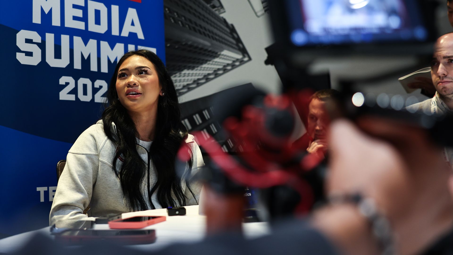 An Asian woman with long dark-brown hair sits at a table strewn with phones and audio recorders in front of a banner reading "Media Summit" 
