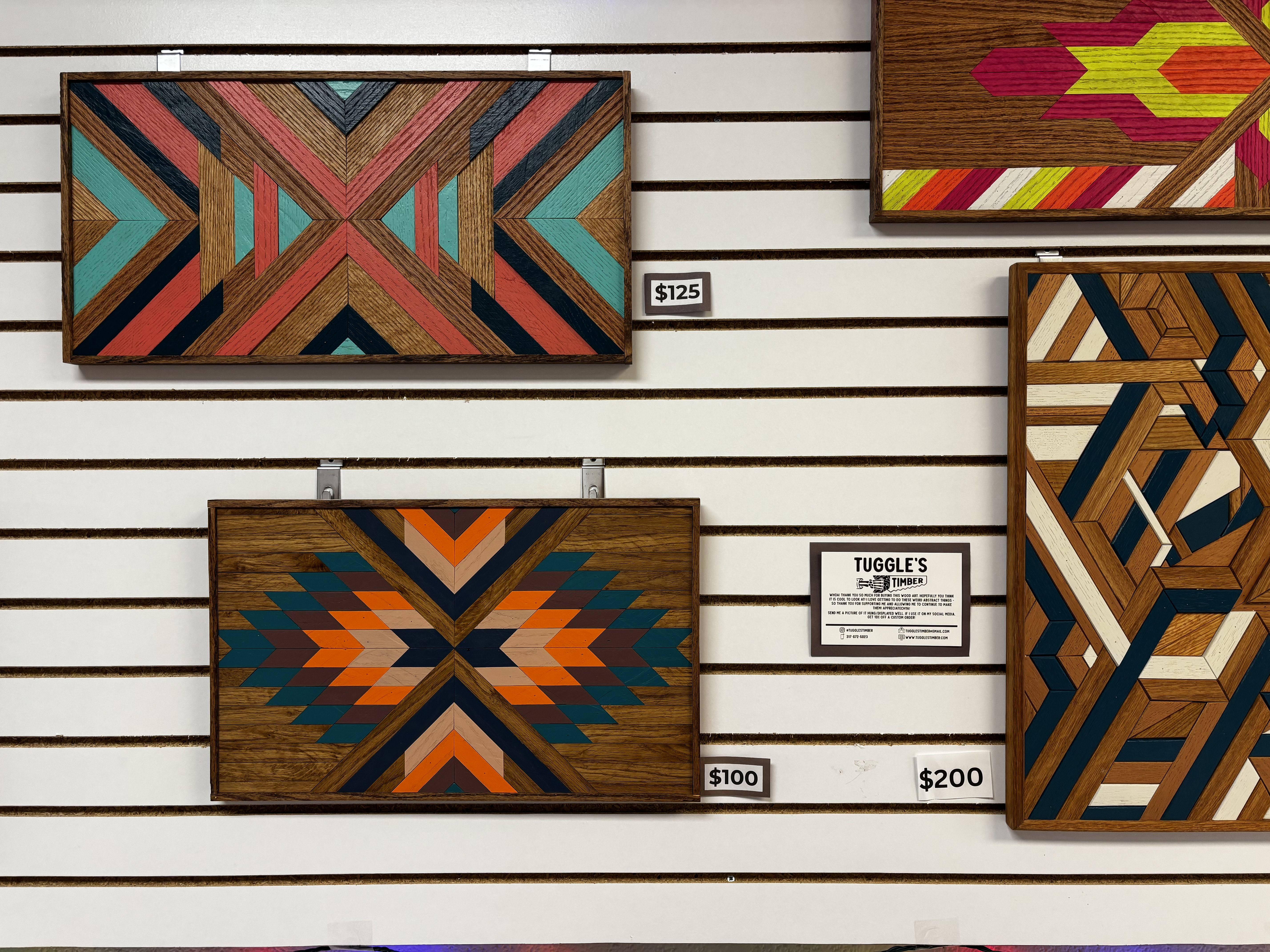 Colorful geometric wooden wall art pieces in shades of orange, teal, black, and natural wood on a white slat wall with price tags $100, $125, and $200, and a small Tuggle's Timber sign.