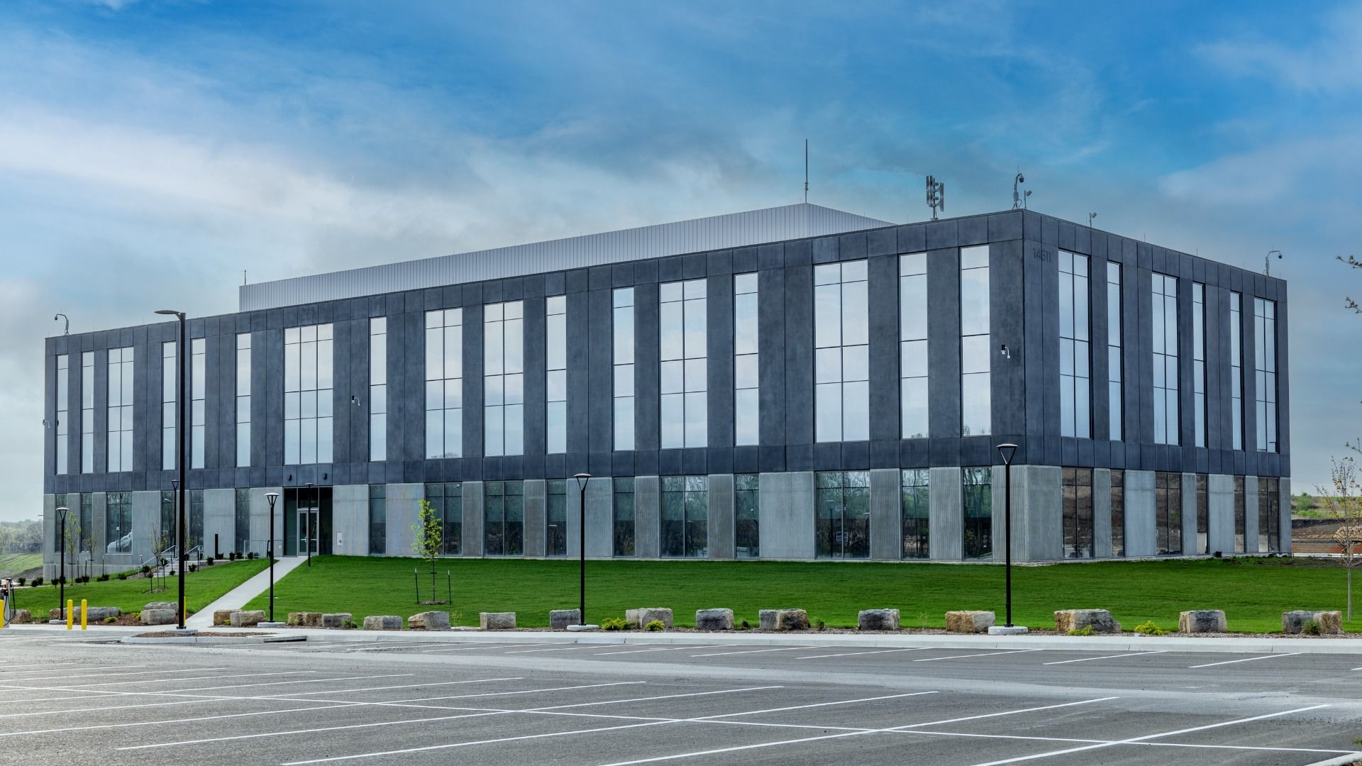 A modern gray office building with tall vertical glass windows sits on a manicured green lawn, with a spacious parking lot in front. Clear blue sky and a few clouds overhead.