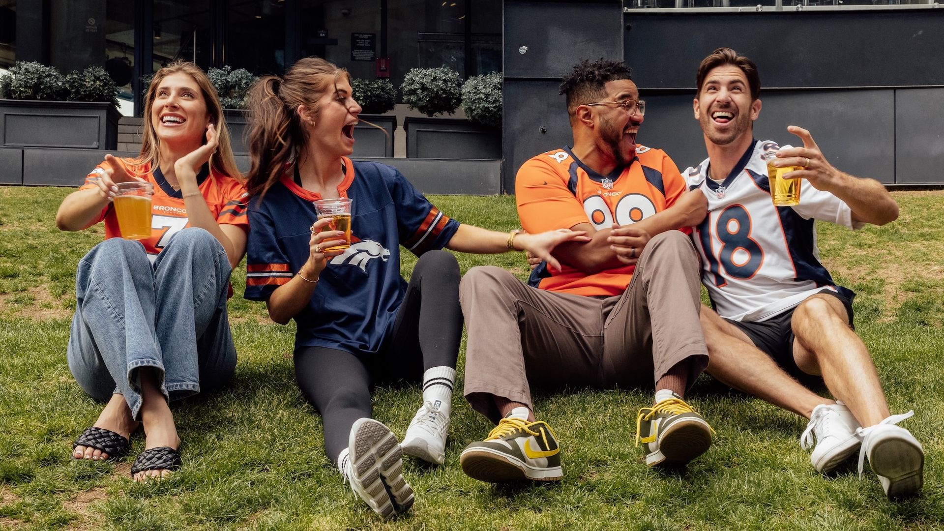Four friends sit on the grass outside a dark building, wearing Denver Broncos jerseys (orange and navy), holding drinks, laughing and cheering as a "Playoffs" banner is visible in the window.