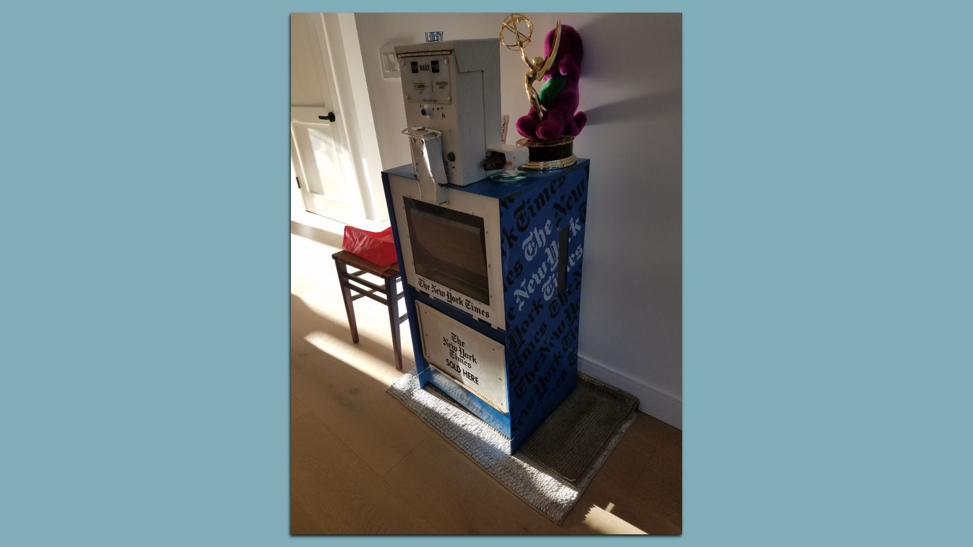 Blue The New York Times newspaper vending machine with a glass front, topped by a gold Emmy statue and a purple plush toy, placed in a sunlit room beside a chair with a red bag.