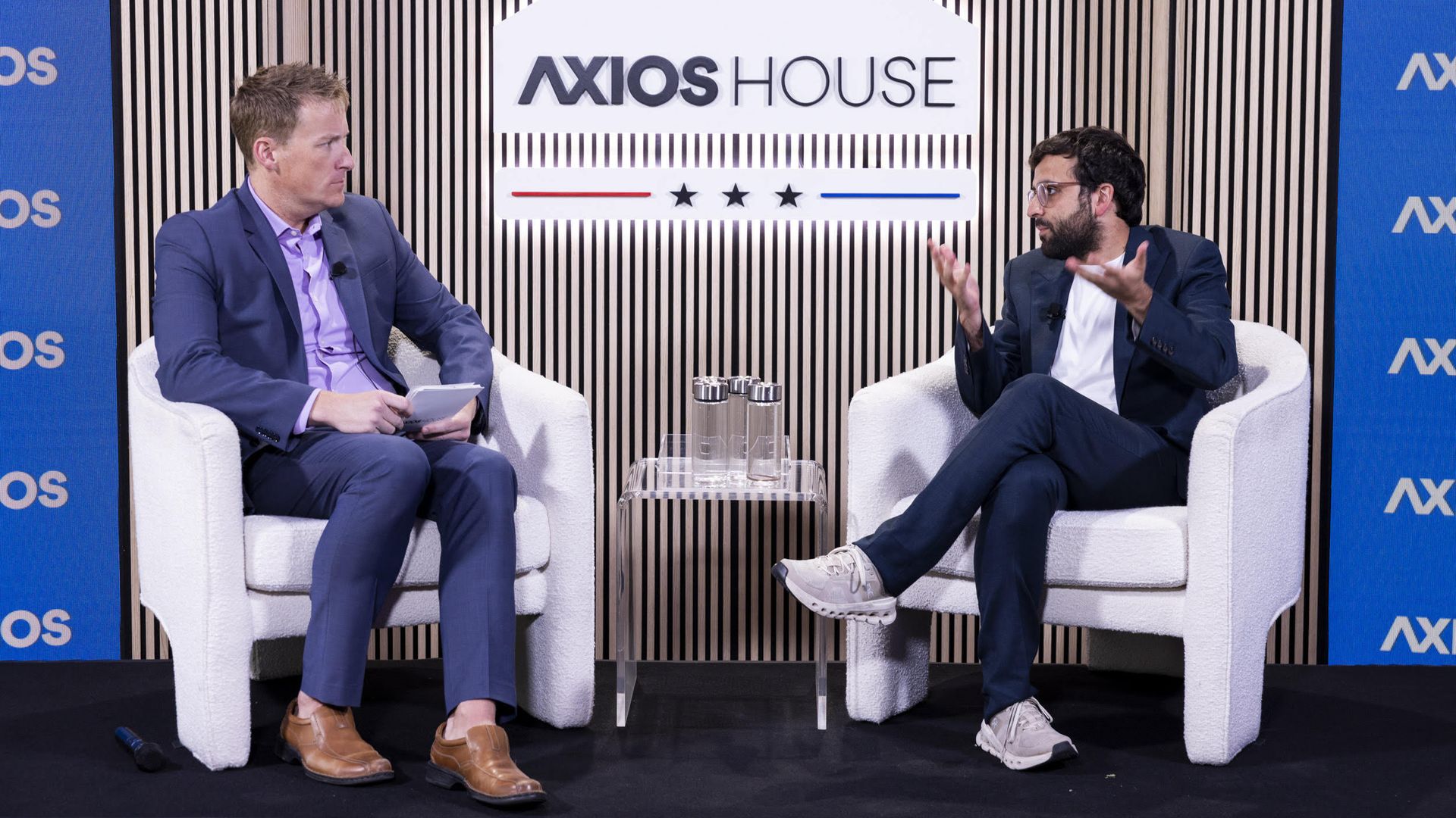 Two men sit in white chairs on a stage with a striped wood backdrop and the large "AXIOS HOUSE" sign. They converse, with a glass table and water between them.
