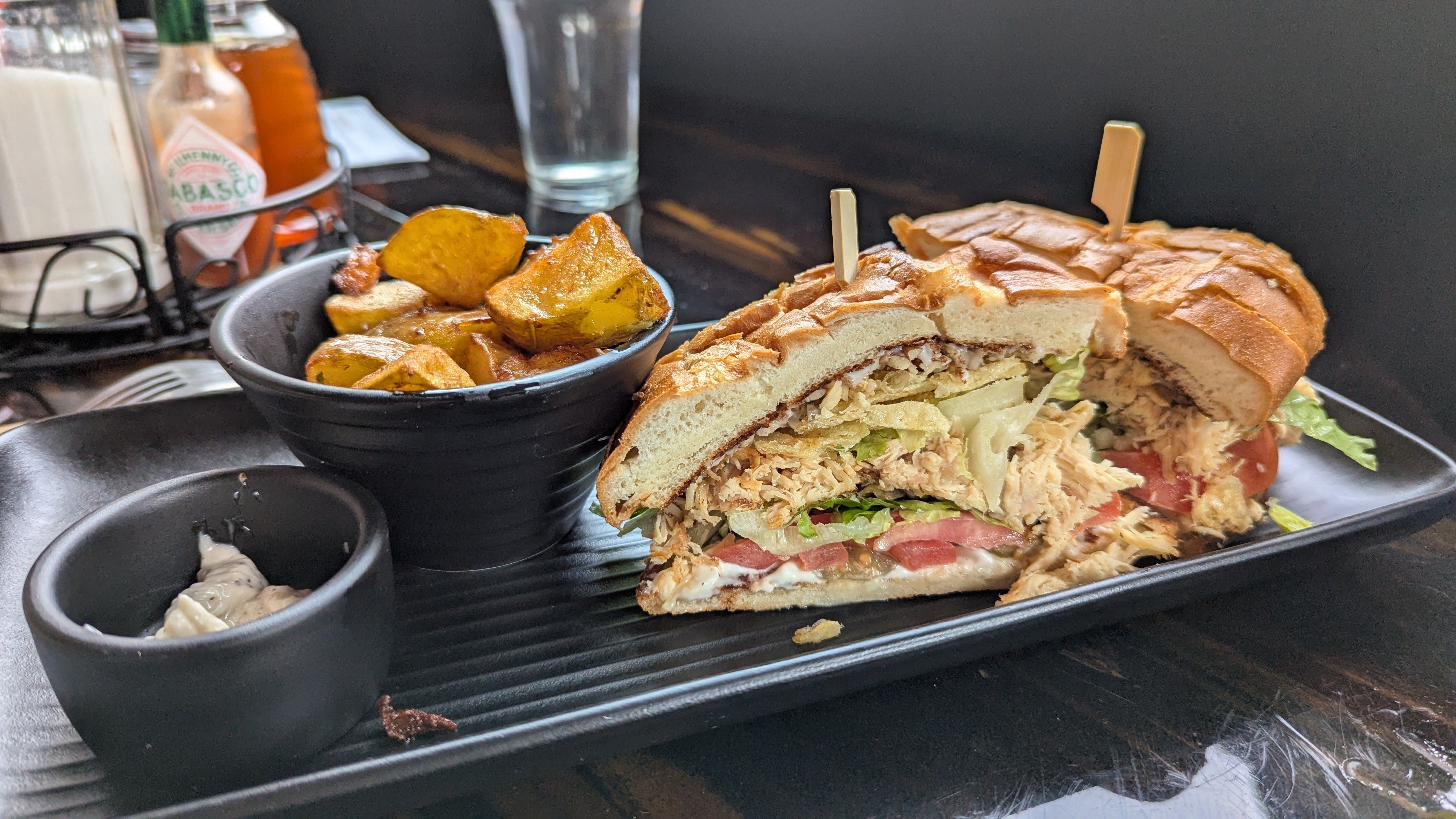 A pulled chicken sandwich on a tray next to a dish of fried potatoes and a bowl of sauce.