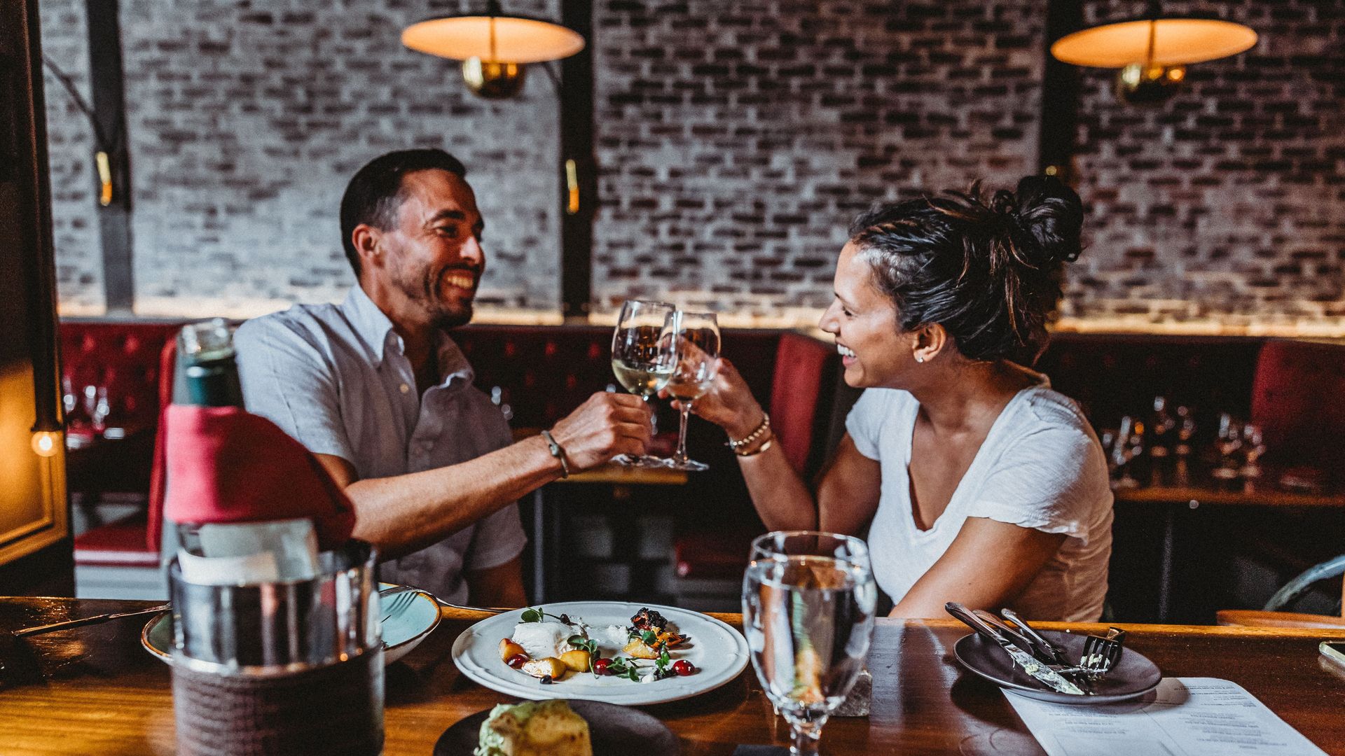 A man and woman smiling and clinking wine glasses across a restaurant table with plates of food, a water glass, and a wine bottle in a holder, with brick wall and red booths behind.