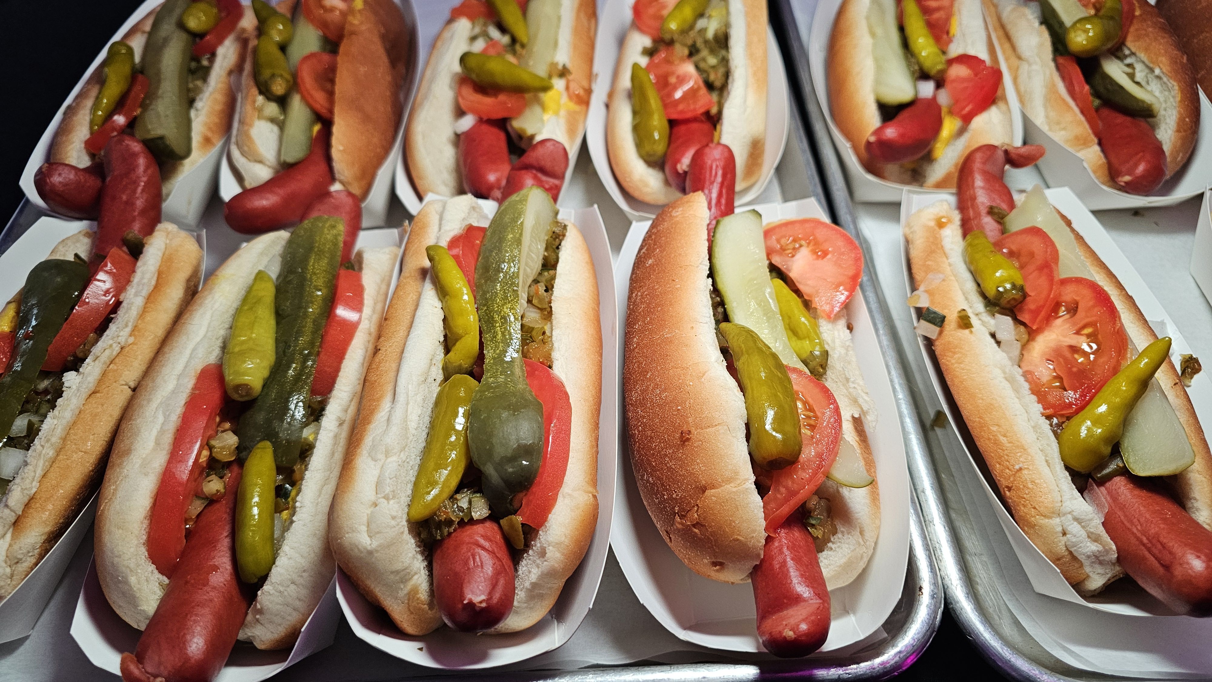 Several hot dogs in buns topped with pickles, tomato slices, onions, and peppers, arranged in white trays on a metal surface.