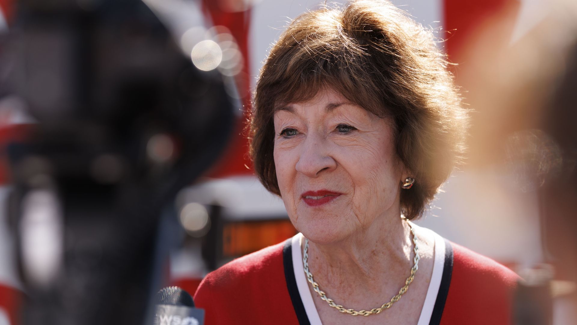 Close-up of an older woman with short brown hair, wearing a red top with white and navy trim and a gold chain. She looks to the side; a microphone is visible in the foreground with a blurred background.