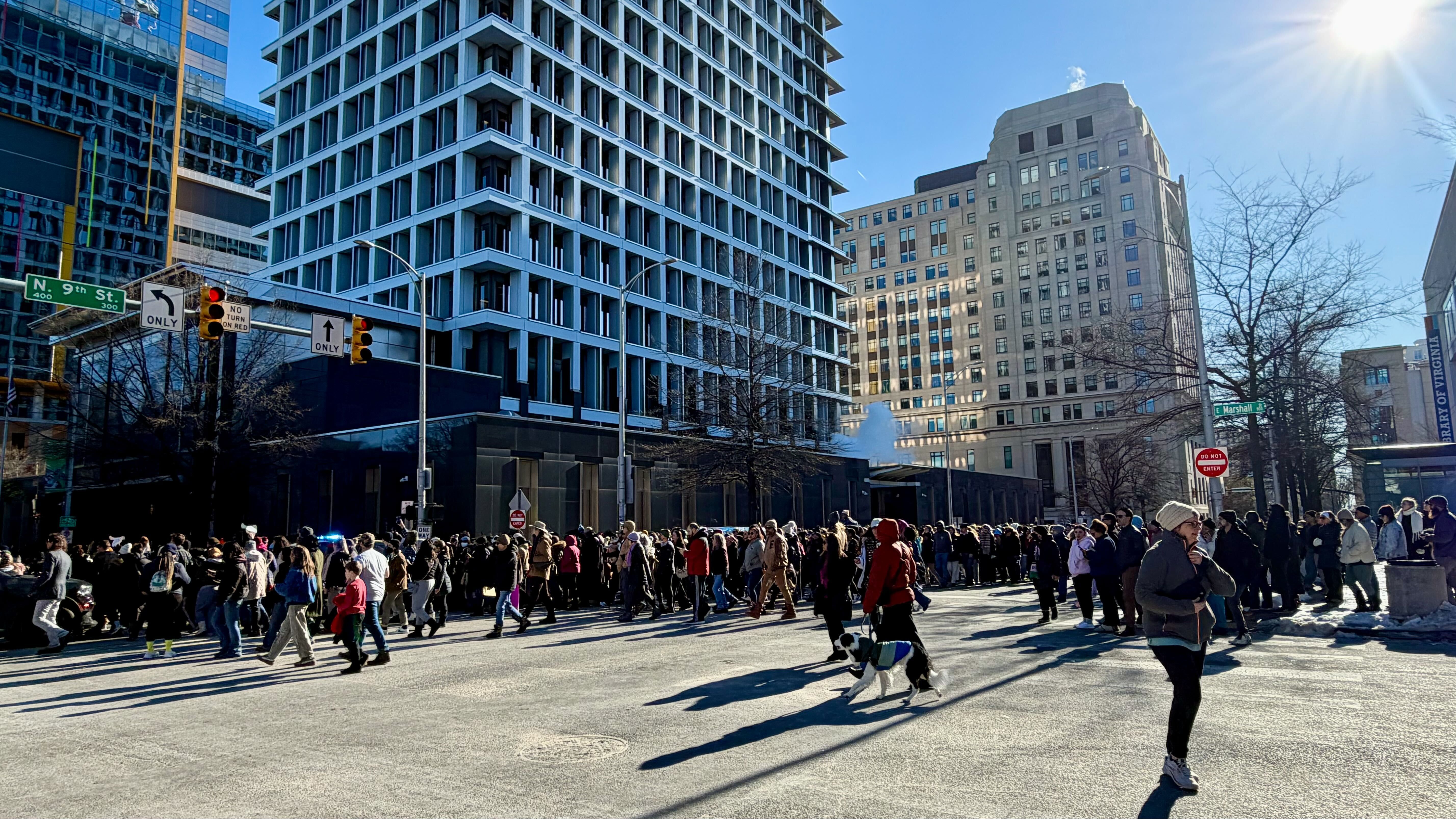 Large crowd of people crossing a sunny city street near tall buildings at N 9th St and E Marshall St, some wearing winter coats and a person walking a dog.