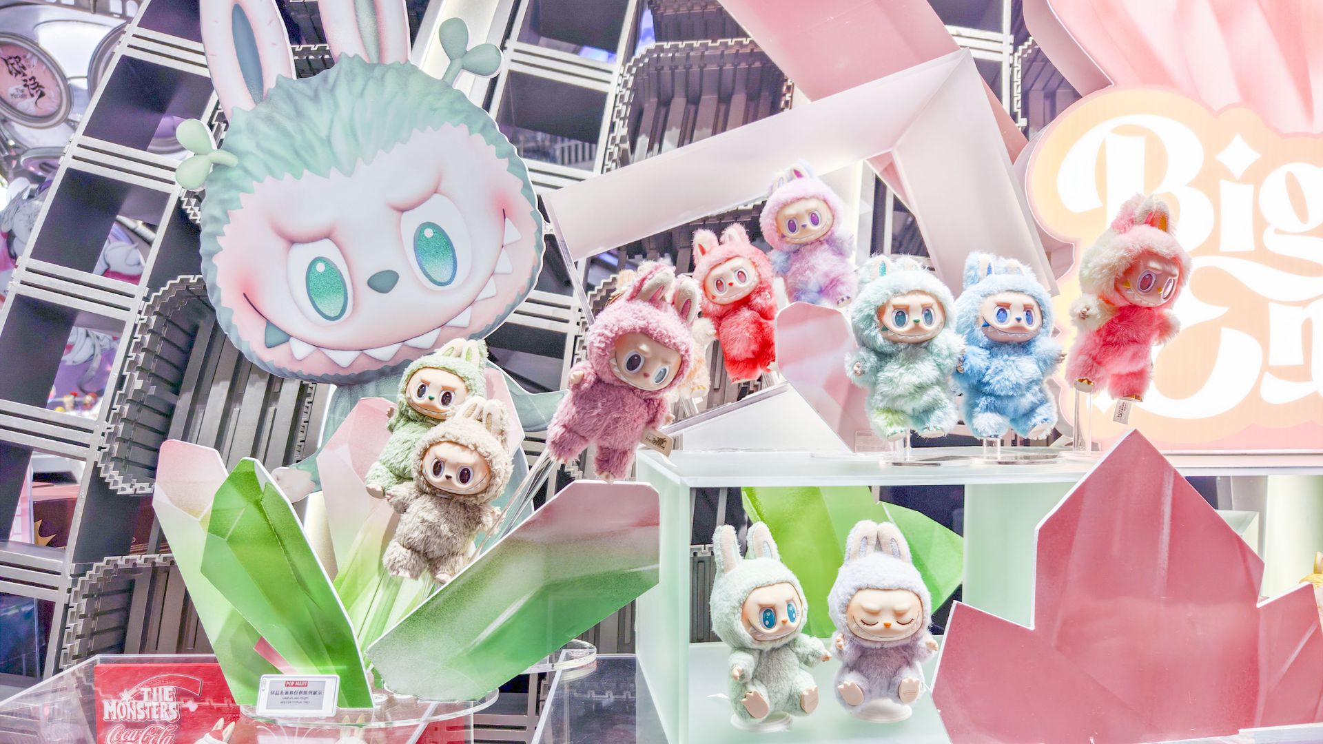 Display of colorful, furry rabbit-eared dolls in pastel shades with large eyes, arranged among green and pink crystal decorations, featuring a large rabbit face backdrop and a sign saying "Big".