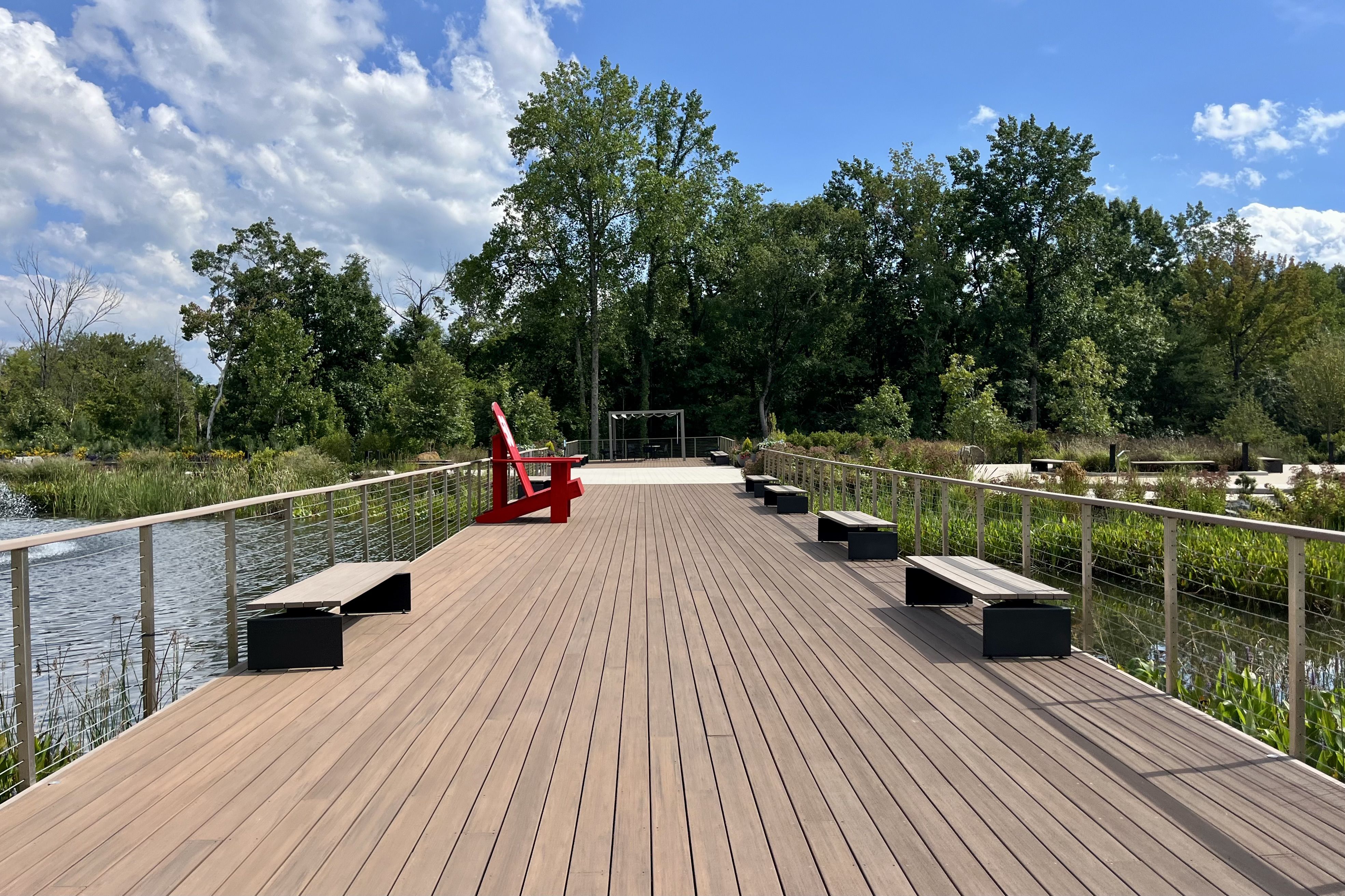 Wide wooden boardwalk with metal railing, small benches on sides, and one large red chair, surrounded by green trees and plants under a partly cloudy blue sky.