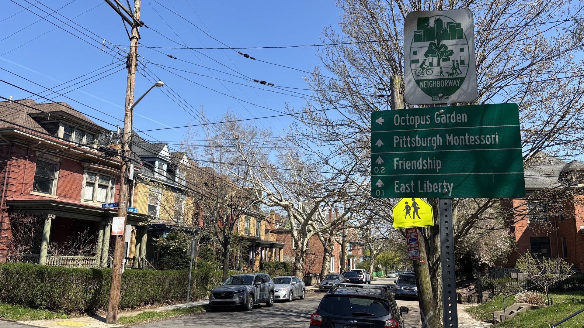 Urban street scene with brick row houses, leafless trees, and a utility pole with wires. A green sign lists Octopus Garden, Pittsburgh Montessori, Friendship, and East Liberty; a yellow pedestrian sign sits below.