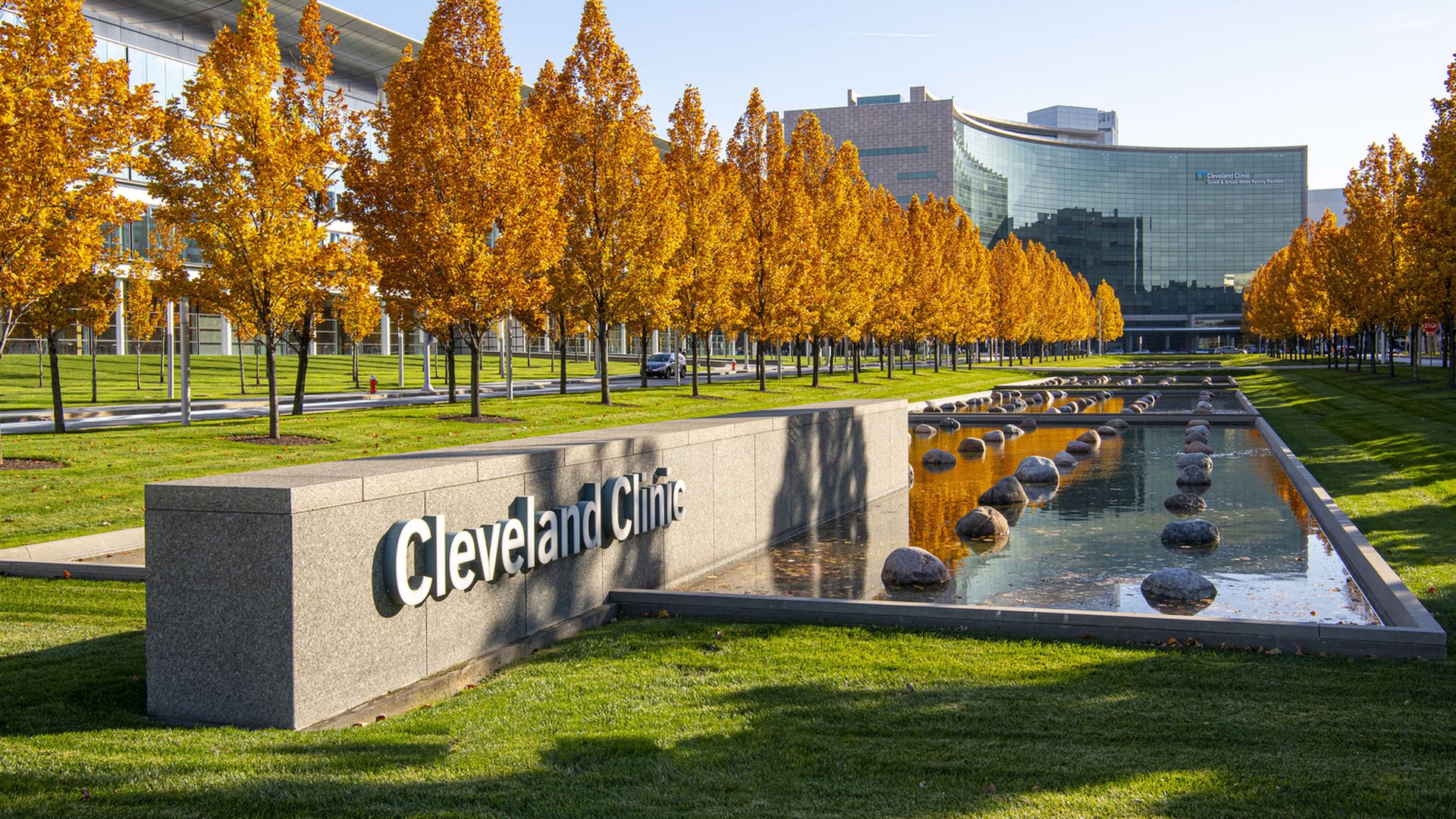 Autumn scene at Cleveland Clinic: row of orange-leaved trees along a reflecting pool with rocks, and a modern curved glass building in the background behind a large Cleveland Clinic sign.
