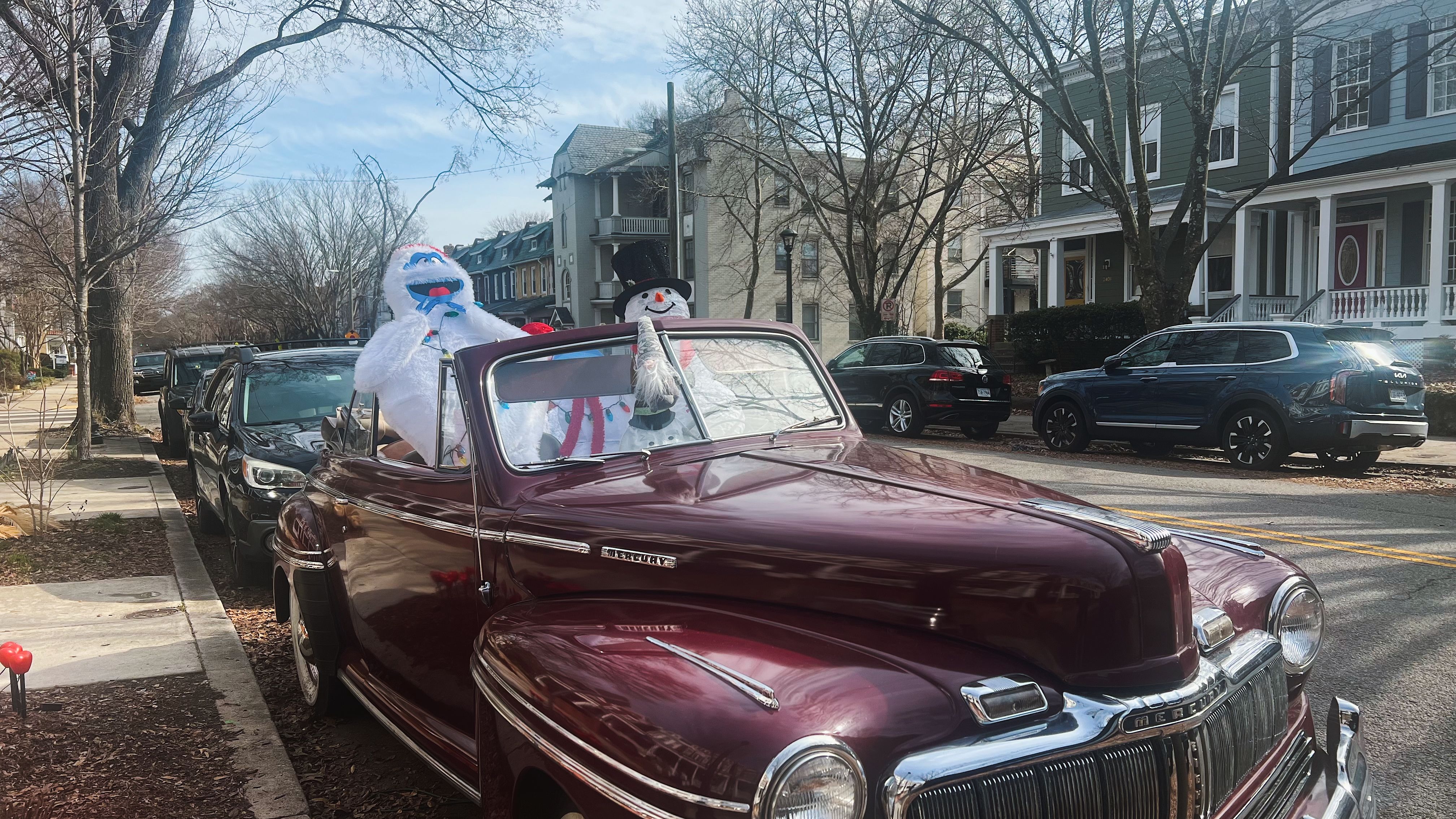 a maroon vintage car on a street with a snowman and bigfoot inside of it