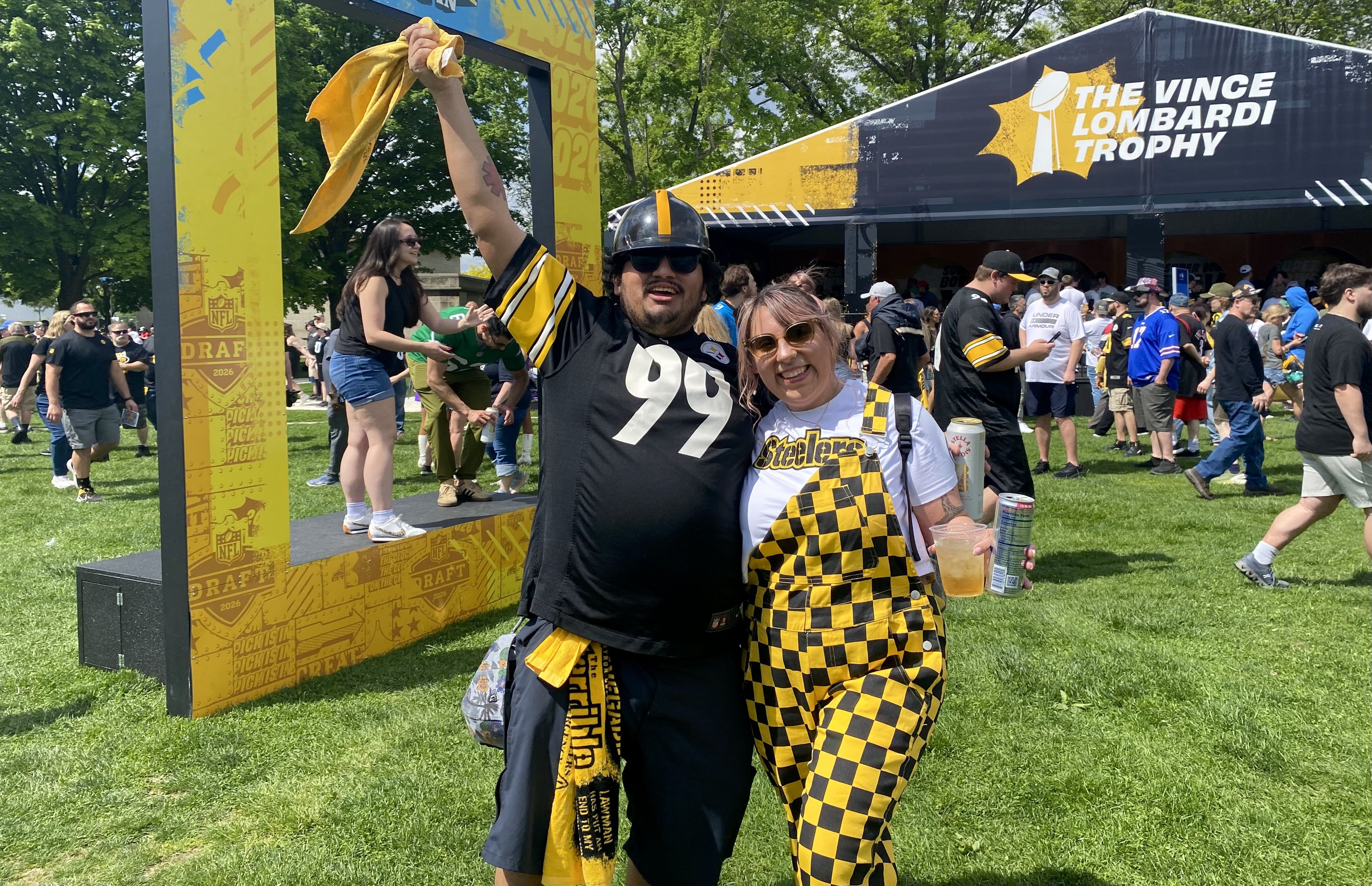 Two Steelers fans in black and yellow outfits pose with a raised scarf, smiling by a yellow NFL Draft frame and a large tent reading "The Vince Lombardi Trophy" at an outdoor event.