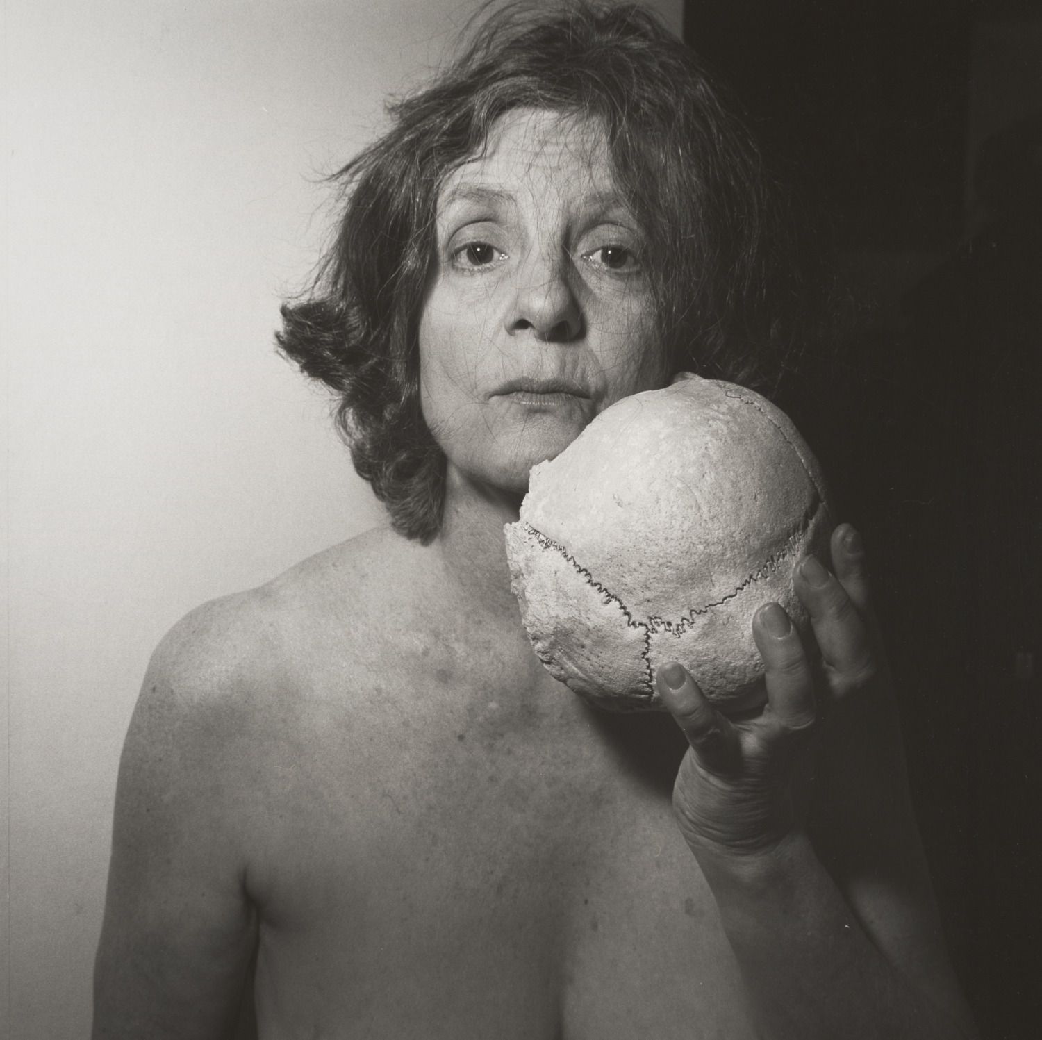 Black and white photo of a topless older woman with tousled hair holding a large skull near her face, looking directly at the camera with a neutral expression.
