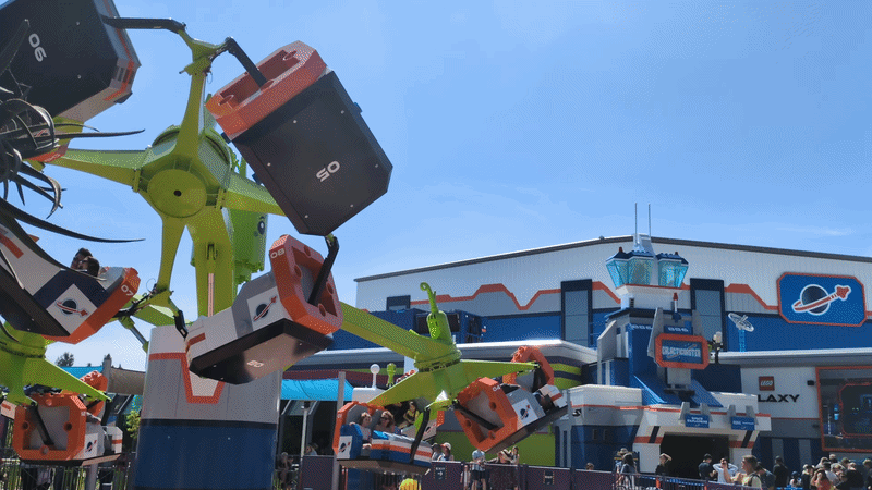 Bright amusement park ride with lime-green arms and orange/black rotating seats; riders visible, blue sky above, and a space-themed building with signage in the background, with a crowd nearby.