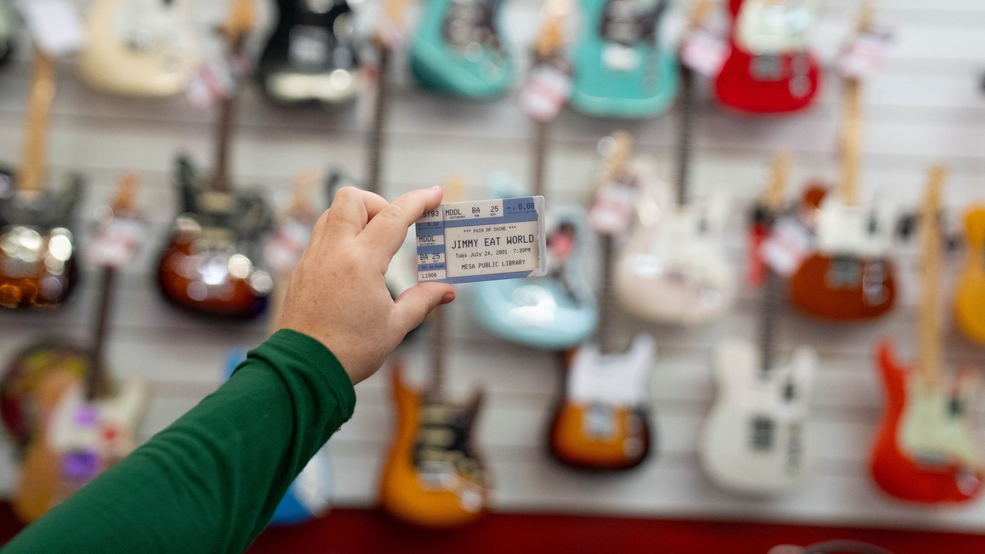 A hand in a green sleeve holds a ticket reading "Jimmy Eat World" in a guitar shop, with a blurred wall of colorful guitars (teal, red, and natural wood) behind.