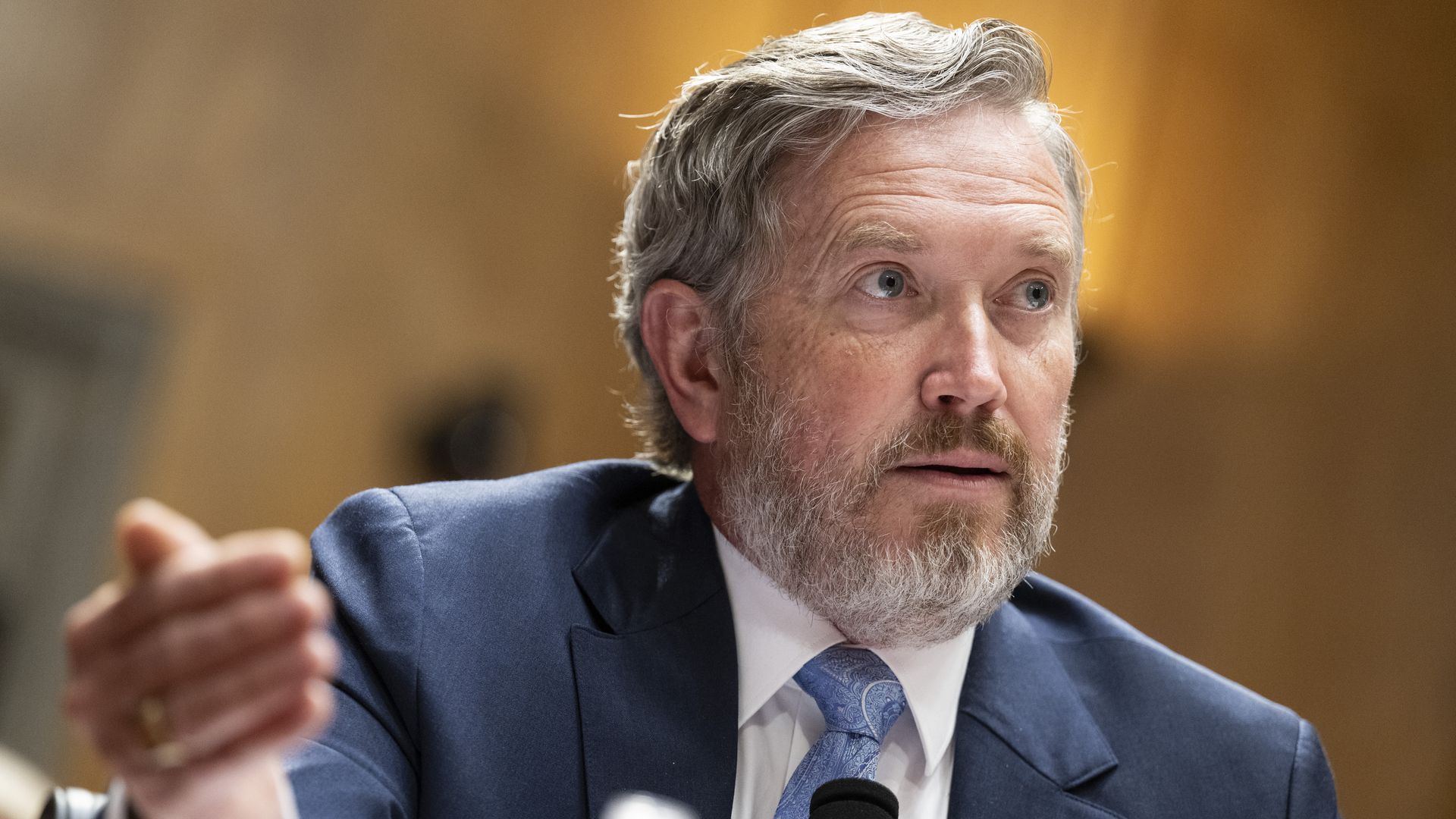 A close-up of a bearded man in a blue suit and light blue tie speaking into a microphone at a panel, gesturing with his left hand; warm wood-toned background.