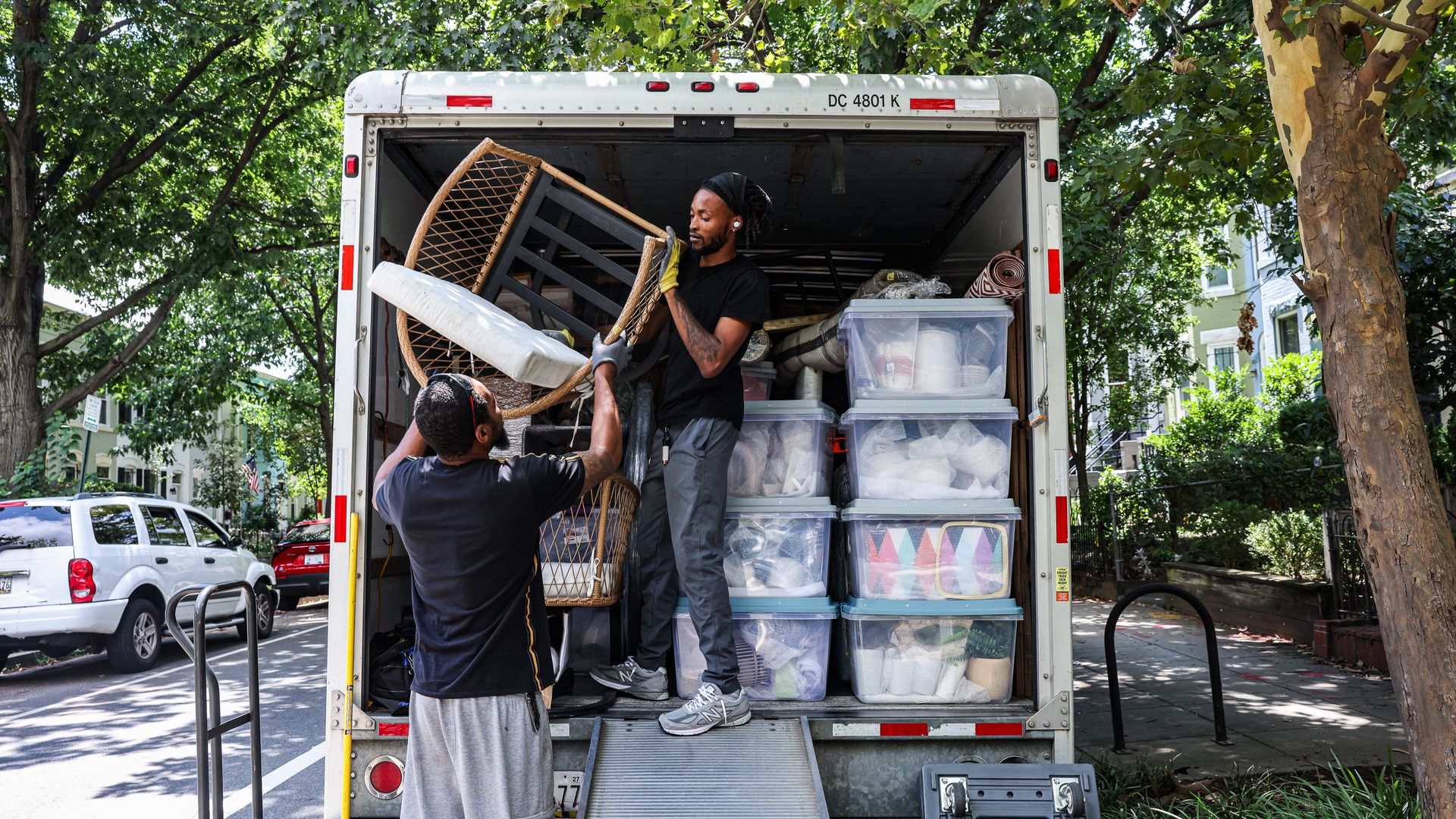 Two men loading a wicker chair into a packed moving truck on a leafy residential street with plastic bins and wrapped items inside the truck.