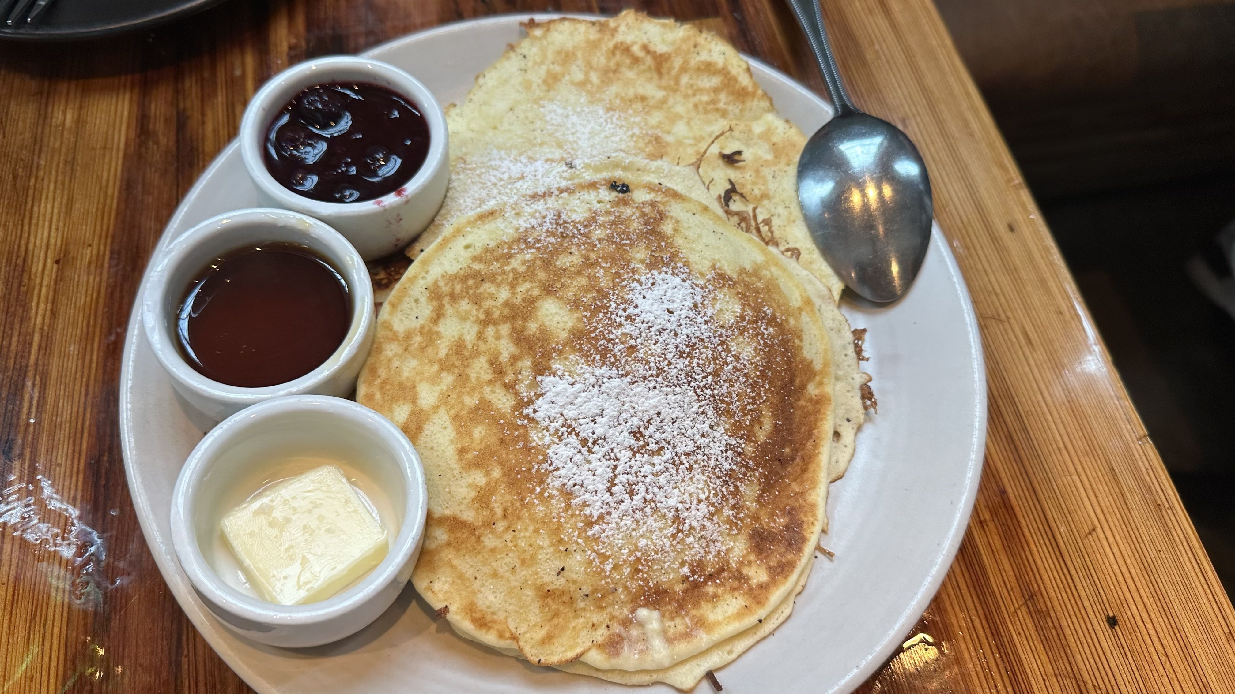 Plate with a stack of golden pancakes dusted with powdered sugar, served with butter, maple syrup, and a blueberry compote in small cups on a wooden table.