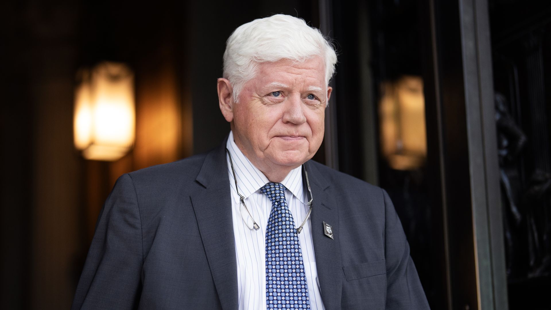 Older man with white hair wearing a dark gray suit, white shirt with thin stripes, blue patterned tie, and glasses hanging around neck, standing near a lighted doorway.